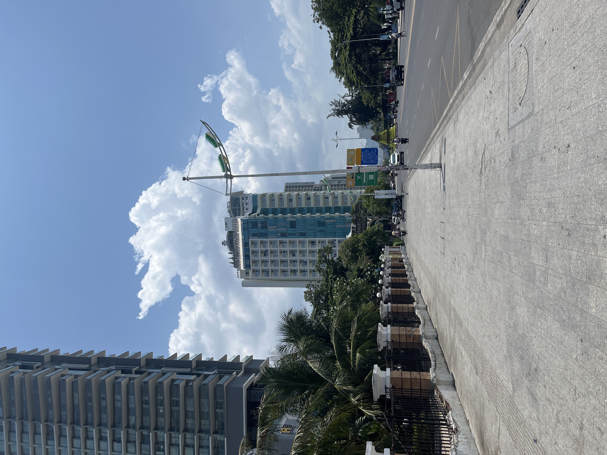The image shows a wide, paved sidewalk next to a street in Nha Trang, Vietnam. The sidewalk has a patterned texture with some visible seams and manhole covers. Along the edge of the sidewalk is a low decorative fence with brick pillars and black metal bars. Beyond the fence, palm trees and other lush greenery are visible.
Several modern, tall buildings line the street. The building on the left is dark grey with numerous balconies and windows. The building in the center is lighter, with a blue-green glass facade and a prominent sign at the top that appears to read "THE LIGHTHOUSE REGAL HOTEL".
The street itself is asphalt with multiple lanes. There are several vehicles on the road, including cars, motorcycles, and scooters. Some pedestrians are visible walking on the sidewalk and crossing the street. A lamppost with two green fixtures is positioned near the center of the image, along the sidewalk. The sky is bright blue with scattered white, puffy clouds. The overall impression is a sunny day in a busy urban environment.