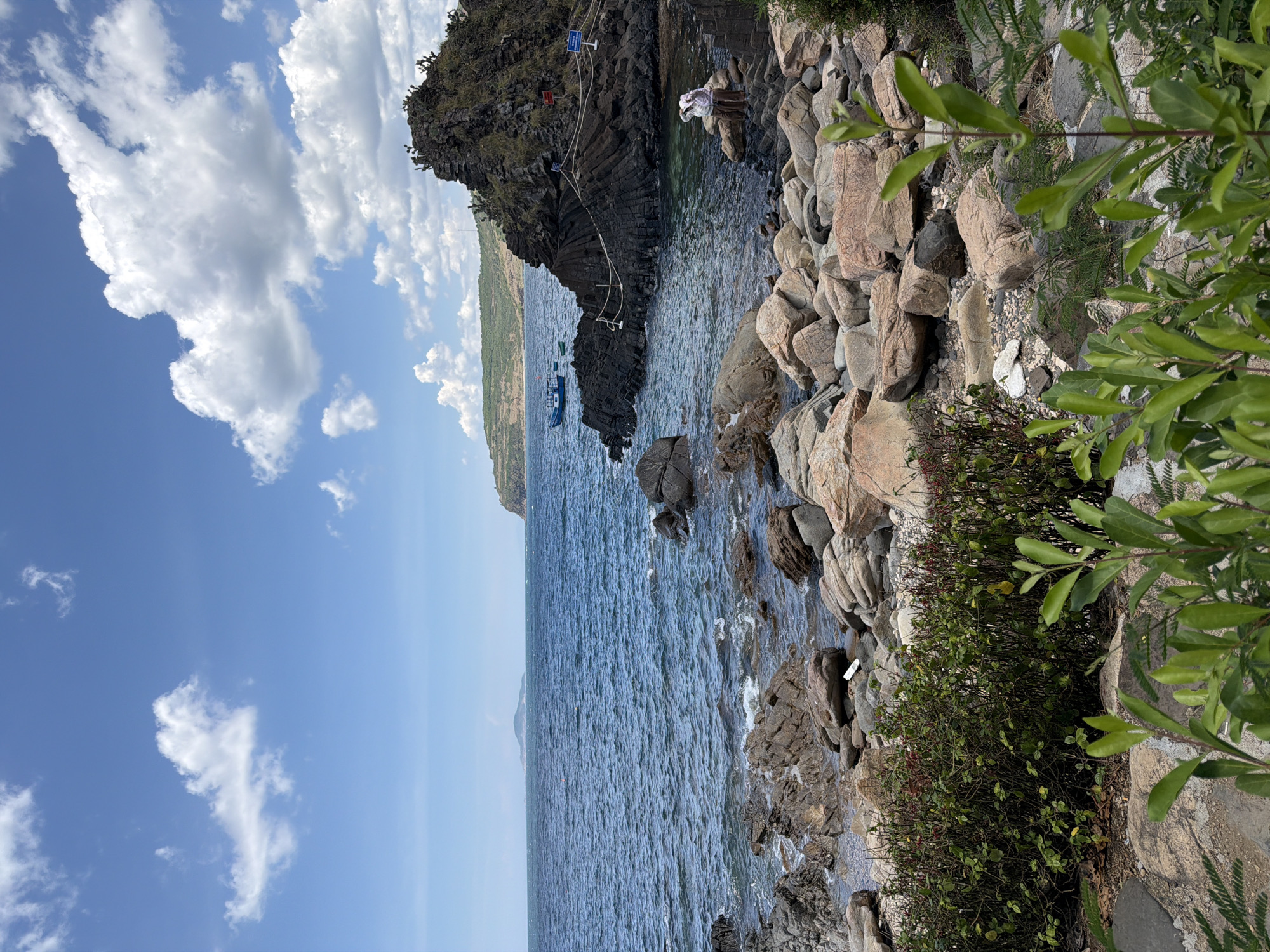 The image depicts a rocky coastline under a blue sky with scattered white clouds. The sea is a deep blue, with small waves lapping against the shore. The shoreline is covered in various sizes of rocks and boulders, with some green vegetation growing between them and in the foreground.
In the mid-ground, a cluster of dark, layered rocks juts out into the sea, forming a small promontory. There are a few small boats anchored in the water near this rock formation. Two people, dressed in light-colored clothing and hats, are visible sitting on the rocks on the right side of the image, seemingly looking out at the sea. A rope barrier with stanchions and a sign are present on the dark rock formation.
The horizon shows a distant island or peninsula with hills under the sky. The overall impression is of a scenic, natural coastal landscape, likely a tourist spot. The location is identified as Phú Lương, Vietnam.