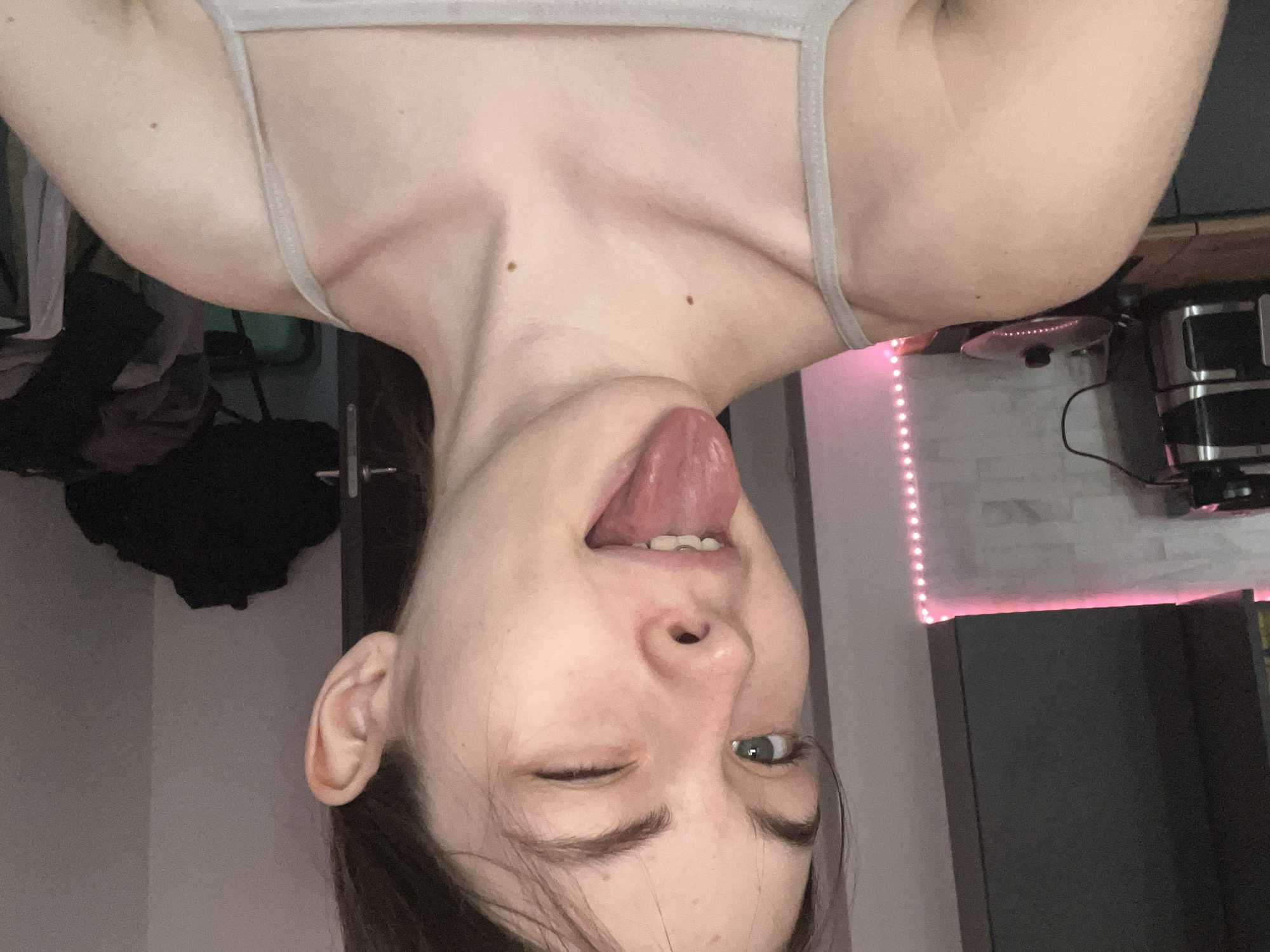 A woman is posing for a selfie, with her head tilted upside down. She is winking with one eye and sticking her tongue out. She is wearing a light-colored tank top. The background is a cluttered room with a closet, desk, and what appears to be a mini fridge. There are LED strip lights emitting a pink glow in the background. The location cannot be determined based on the provided image.