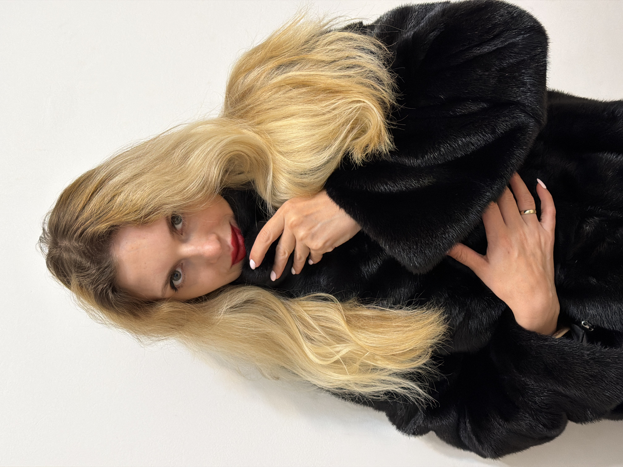 A medium close-up shot captures a woman with long, flowing blonde hair and red lipstick, wearing a luxurious black fur coat. The image is oriented diagonally, with the woman's head positioned towards the lower left corner and her body extending upwards and right. She appears to be lying down or leaning back against a plain white background, looking directly at the viewer with a calm, neutral expression.
Her hands are visible against the dark fur of her coat. One hand is placed gently on her arm, while the other rests near her chest, both displaying neat, light pink manicured nails. A simple, slender gold band ring is visible on the ring finger of her right hand. The black fur coat has a soft, dense texture, and her blonde hair cascades around her head and over the coat, creating a strong contrast against the dark material and the white background. No specific location cues are visible within the frame, but the provided context for the image is Budva, Montenegro. There is no visible text in the image.