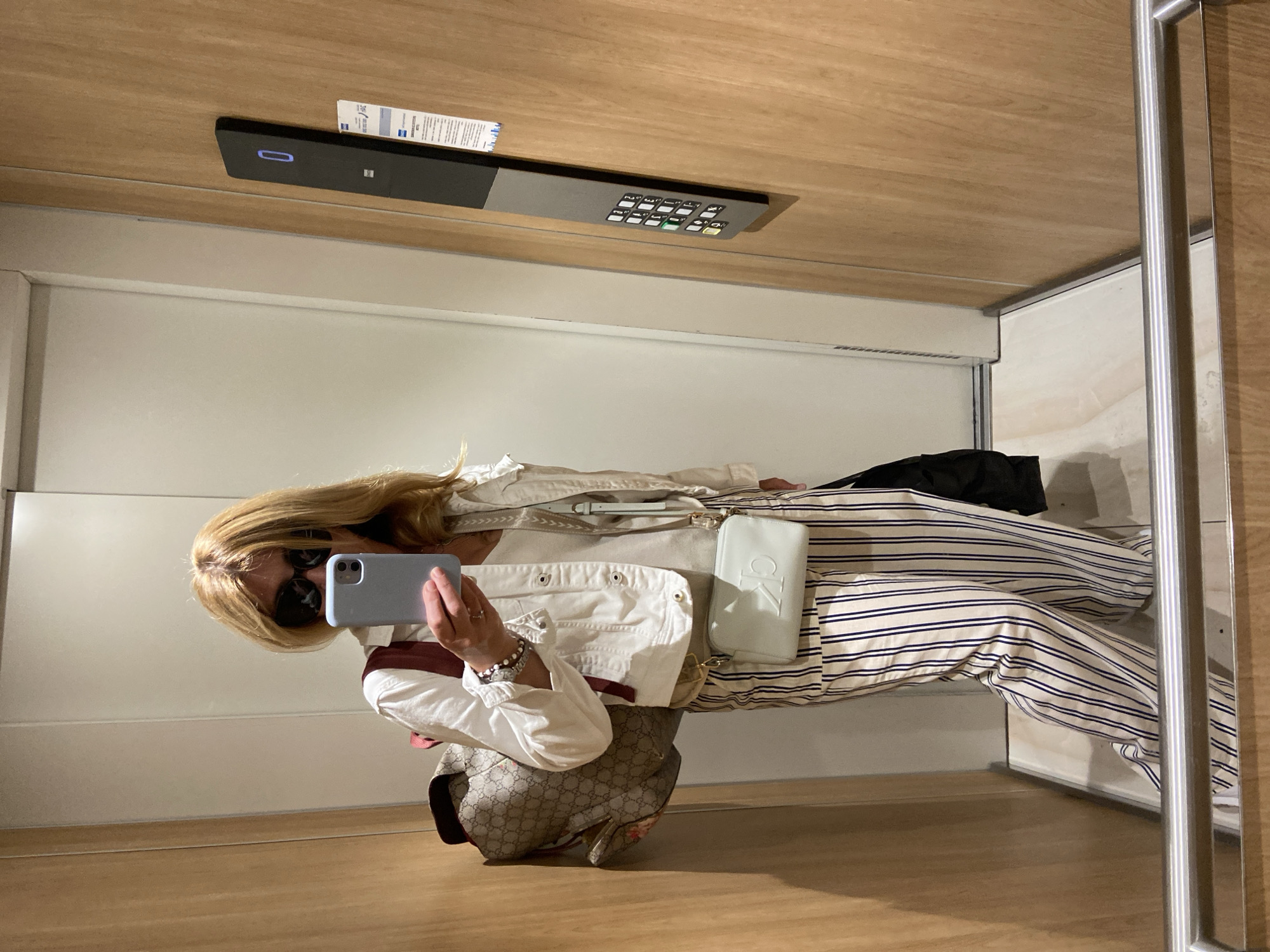 A person is captured in a mirror selfie within an indoor setting, likely a hotel room in Budva, Montenegro. The individual, appearing female, has long blonde hair and wears sunglasses. They are dressed in a white jacket or shirt over horizontally striped pants (navy and white), and hold a light blue/grey smartphone (resembling an iPhone) to take the photo.
Visible accessories include bracelets on the wrist, a light blue or white Calvin Klein ("CK") clutch bag, and another light-colored bag with a patterned strap. A larger bag with a distinctive Gucci monogram pattern rests on the floor next to the person.
The room features wooden surfaces for the floor and ceiling, and white walls. On the wall/ceiling, a black rectangular smart device with a blue indicator light is visible. A small white paper, possibly a key card or informational card with text including "ZSC" and other indistinct words, is inserted into this device. Adjacent to it, a grey control panel with several buttons and symbols, likely a keypad for room functions, is present. A metallic frame or handle is visible on the right side, suggesting a door or wardrobe. The overall scene depicts a casual moment in a modern interior.