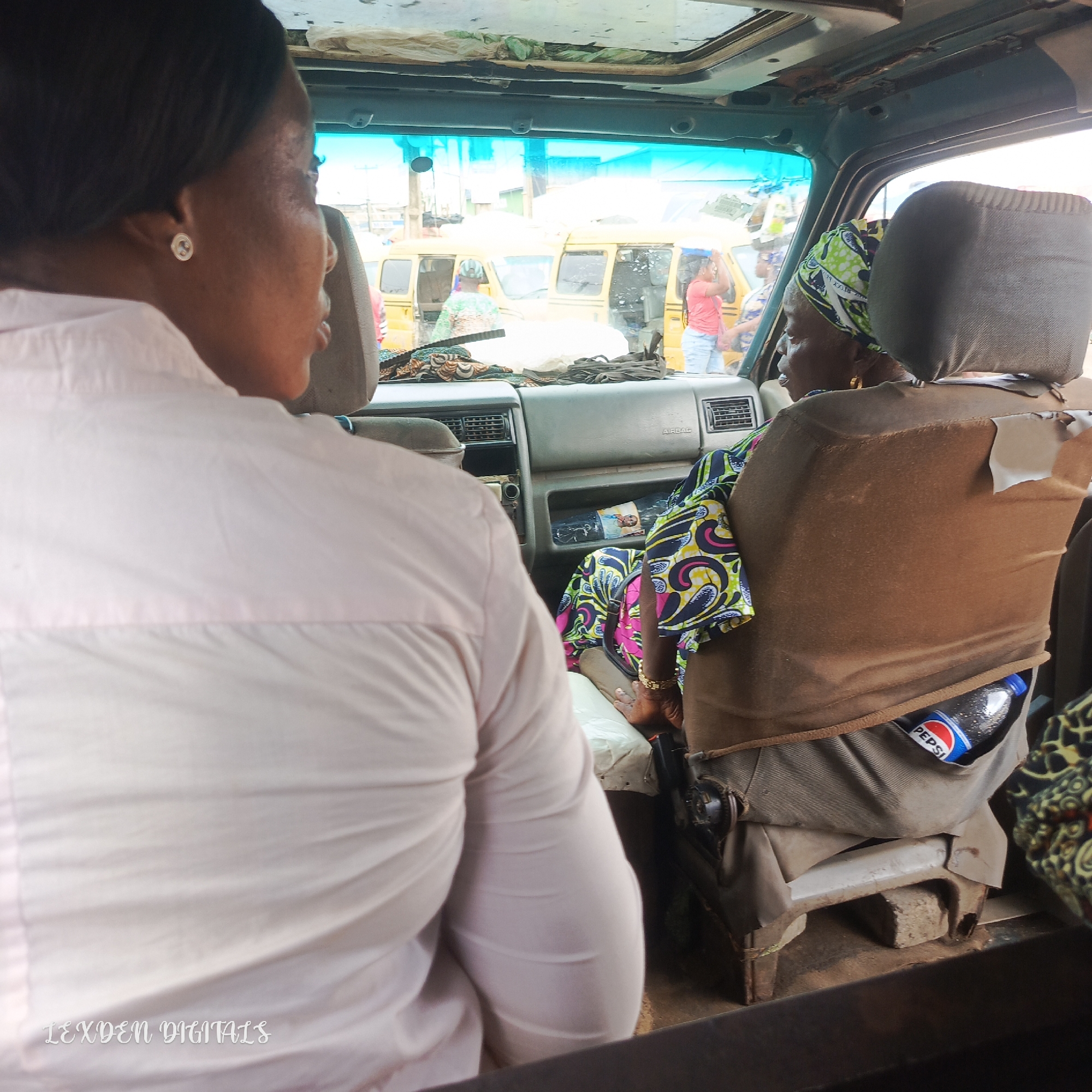 The image shows the interior of a vehicle, likely a commercial bus, with two women visible. The woman in the foreground is wearing a light pink shirt and her back is to the camera, though her profile is partially visible. She has dark hair and is wearing a small silver earring. The second woman is seated to her right, also facing away from the camera but with her head turned slightly to the side. She is wearing a colorful headscarf with green, yellow, blue, and black patterns, and a matching patterned dress. She has gold jewelry on her wrist and is wearing earrings.
The vehicle appears to be in motion or stopped in a busy area, as evidenced by the yellow commercial vehicles visible through the windshield. The dashboard of the vehicle is visible, with air vents and a compartment. A bottle of Pepsi is tucked into a pocket on the back of the second woman's seat. The interior of the bus shows signs of wear and tear, with some damage to the seat covers and ceiling.
Outside the vehicle, there are glimpses of people and other vehicles, suggesting a bustling urban environment. The lighting indicates it is daytime. The overall impression is of a common scene in transportation within a Nigerian city like Egan. The text "LEXDEN DIGITALS" is visible in the lower left corner of the image.