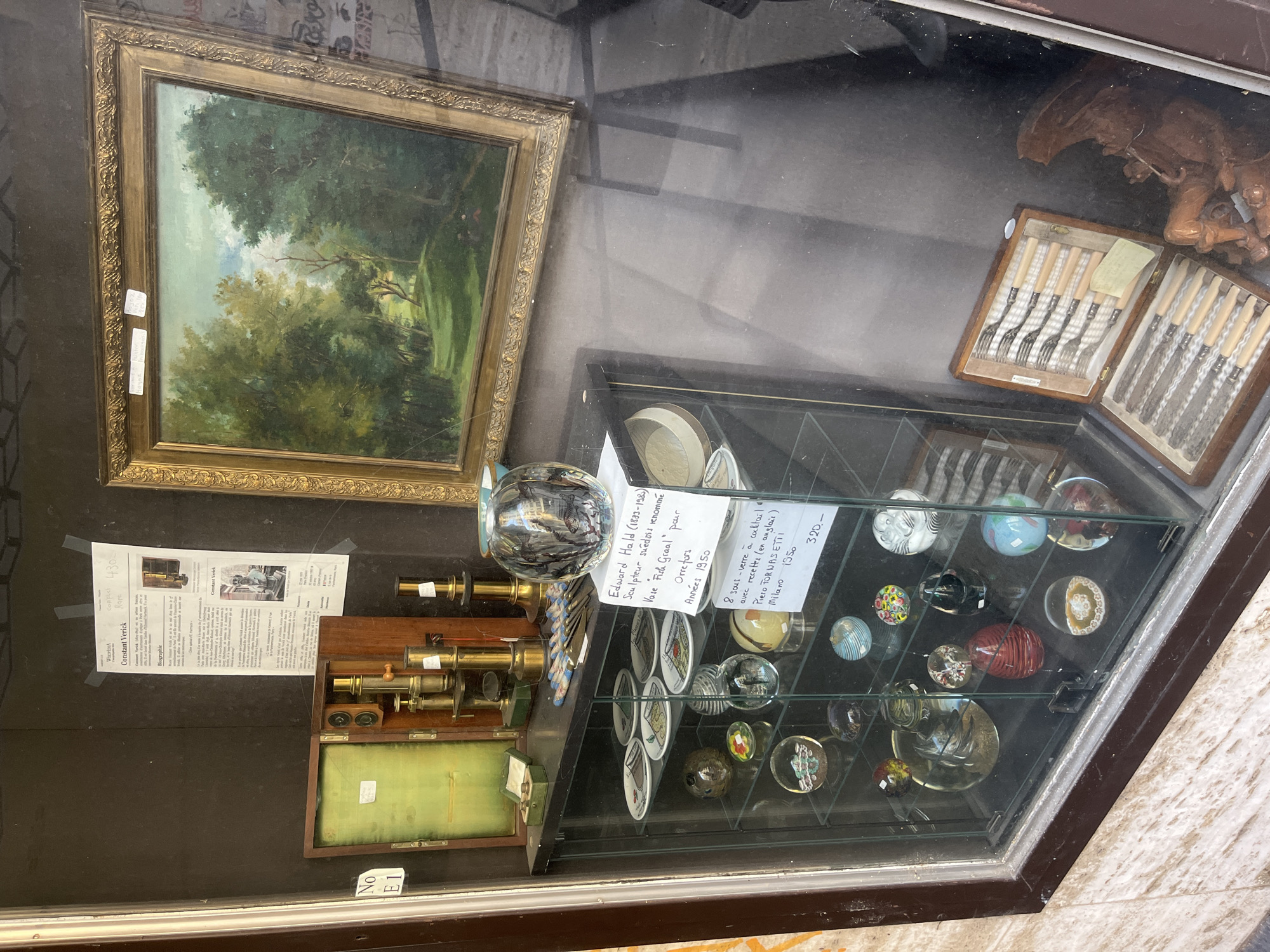 The image shows a display window, possibly a shop, in Vevey, Switzerland. The display features a variety of antique and collectible items. On the top shelf, there is an open wooden box containing a brass microscope and other scientific instruments. To the right of the microscope, a framed landscape painting hangs.
Below the top shelf, a glass-fronted cabinet showcases more items. The upper section of this cabinet holds a collection of small plates and a decorative glass vase. A handwritten card on the cabinet reads "Edward Hald (1893-1982) Sculpteur suédois renommé Vase 'Fish Graal' pour Orrefors Années 1950".
The lower section of the glass cabinet is filled with numerous glass paperweights, some of which appear to be marbles with intricate designs inside. Another handwritten card on this section states "8 sous-verre à cocktail avec recettes (en anglais) Piero Fornasetti Milano 1950 320-".
To the right of the glass cabinet, a wooden box is open, revealing a set of antique cutlery, likely forks and knives, with light-colored handles. Next to the cutlery, a dark brown wooden carving of a winged creature, possibly a gargoyle, is visible. The overall impression is of a curated collection of decorative and scientific antiques presented in a shop window. The location in Vevey, Switzerland, is inferred from the presence of French text on one of the informational cards.