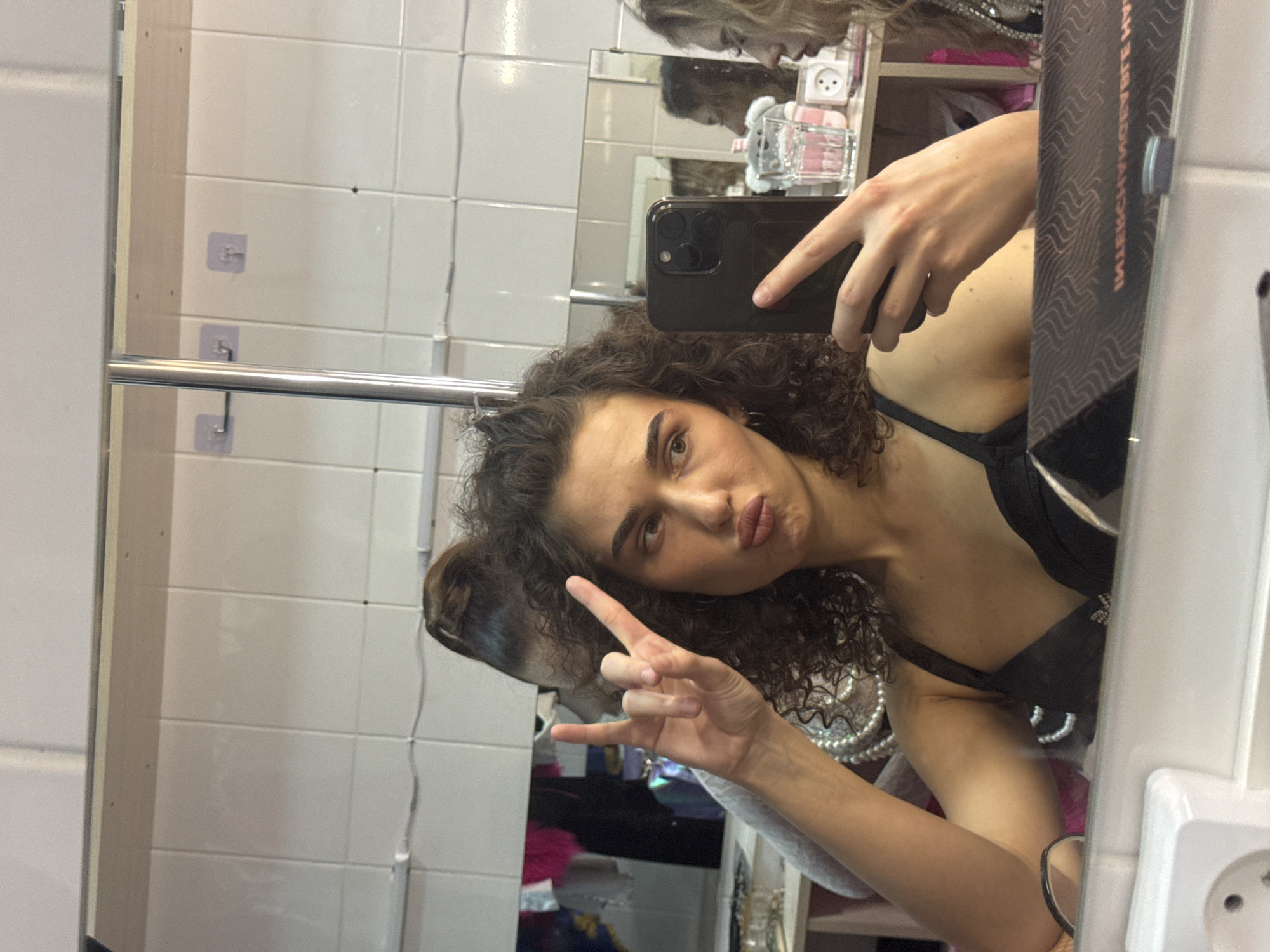 The image is a selfie taken by a young woman with curly brown hair in a black top, posing in front of a mirror. She is making a "rock on" hand gesture with her left hand and holding a smartphone in her right hand, capturing the reflection. The background appears to be a dressing room or backstage area, with tiled walls, shelving, and various items, including what look like theatrical costumes or accessories. There are also reflections of other people and objects in the mirrors. The image does not contain information that allows for identifying the specific location in the city of Kazan, Russia.