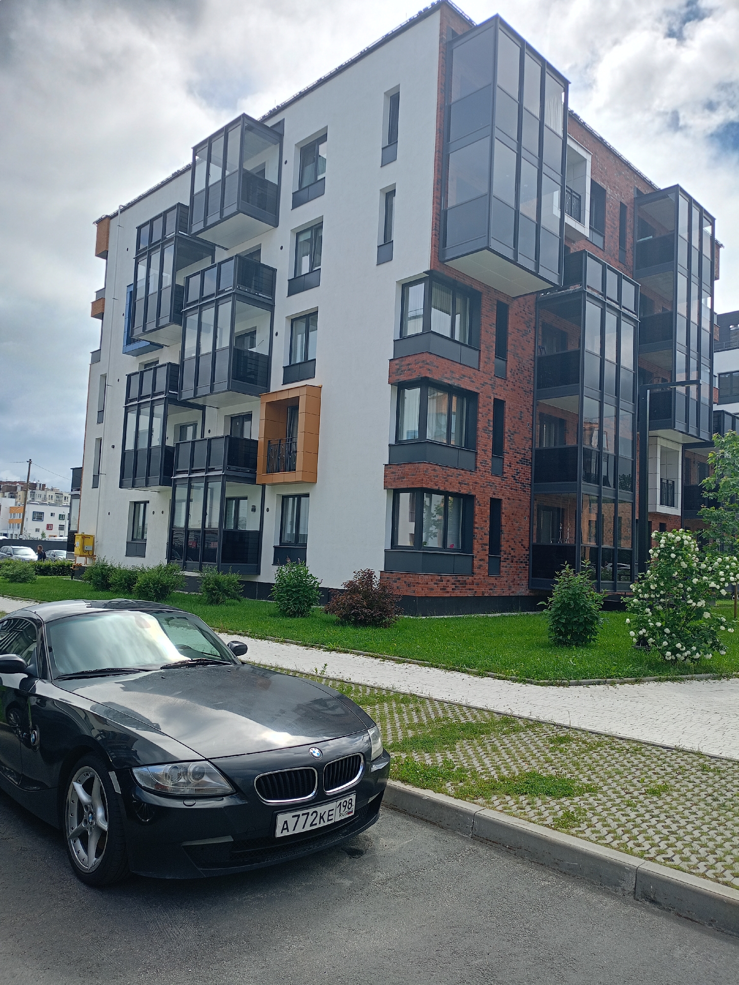 In an urban setting, a black BMW Z4 is parked on an asphalt road beside a concrete curb and a cobblestone pathway with grass growing through the pavers. Behind the pathway is a well-maintained green lawn with manicured bushes and a blooming white bush. Dominating the background is a modern multi-story apartment building with white and red brick facades. The building features numerous glass-enclosed balconies that extend outwards from the structure. The sky above is overcast with scattered clouds.