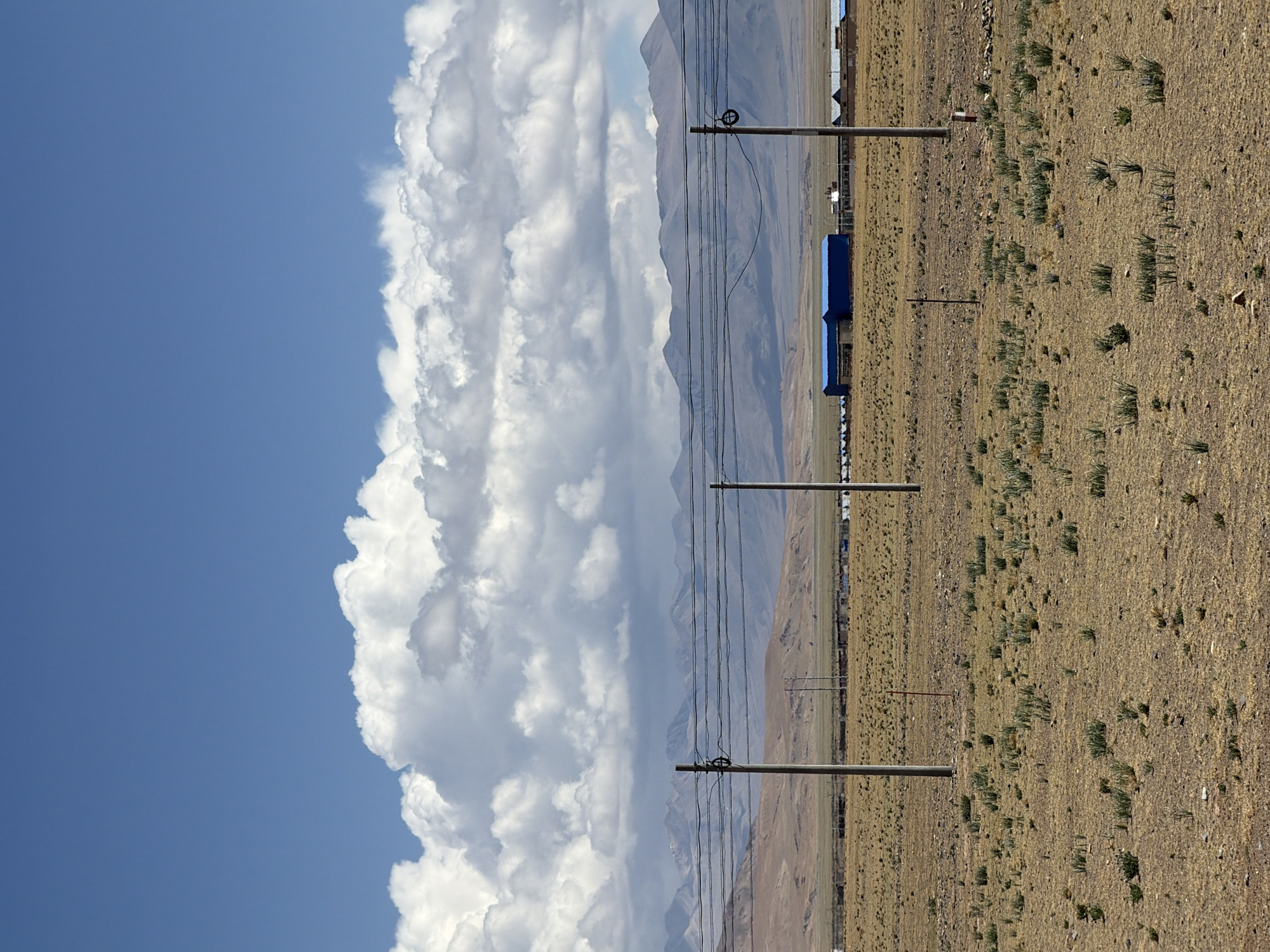 The image captures a vast, open landscape under a clear blue sky, dominated by natural and distant man-made elements. The upper portion of the frame features a bright blue sky, with large, white, fluffy cumulus clouds prominent on the right side, suggesting good weather.
In the middle ground, a hazy, undulating mountain range stretches across the horizon, providing a distant backdrop to the scene. The foreground and mid-ground consist of dry, golden-brown ground, indicative of an arid or semi-arid steppe or plain. This ground is sparsely covered with small, green, reedy clumps of vegetation.
Several utility poles, likely concrete, stand at intervals across the scene, supporting multiple parallel power lines that run horizontally from the left to the right of the frame. In the far distance on the right, near the base of the mountains, a few small buildings or structures are visible, some appearing to have blue roofs.
The lighting suggests it is bright daytime, likely late morning or early afternoon. There is no visible human or animal activity. No text is discernible in the image. The scene evokes a sense of expansiveness and quiet solitude, characteristic of a remote, high-altitude region like Mainpu township, China.