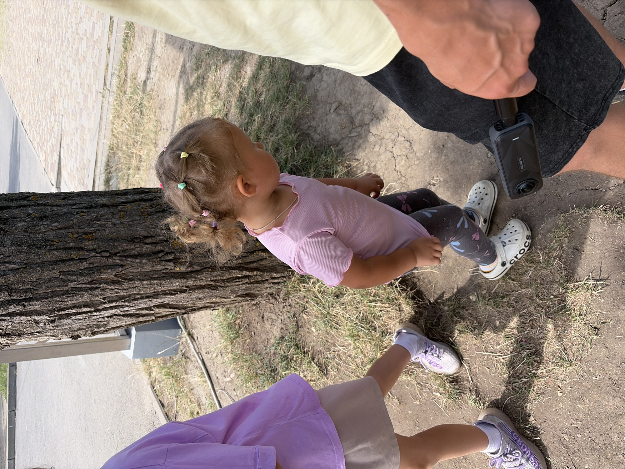 The image depicts an outdoor scene with three individuals. In the foreground, a person's hand, with visible veins and skin texture, is holding a black Insta360 X3 camera attached to a black grip. The camera is pointed downwards towards the ground. To the left, a young child wearing a purple shirt and a light brown skirt is partially visible, with their legs and feet in the frame. The child is wearing white socks and purple sneakers with laces. In the center and to the right, a younger child, a toddler, is wearing a pink striped t-shirt and grey leggings with a bow pattern. The toddler has blonde hair styled with multiple colorful hair ties. The child is wearing white Crocs. A large tree trunk is prominently located in the background, with its rough bark texture visible. The ground is covered with dirt, dry grass, and some small stones. In the upper background, a paved walkway with a cobblestone pattern can be seen, adjacent to a road. A grey concrete structure is also visible near the tree. The scene suggests a casual outdoor activity, possibly a park or a street with a sidewalk. The location is identified as Rostov-on-Don, Russia.