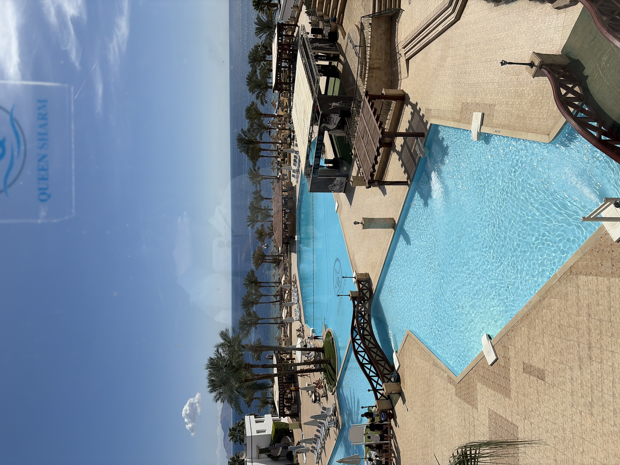The image displays an elevated perspective of the Queen Sharm Resort in Sharm El Sheikh, Egypt, under a clear blue sky with minimal cloud cover. The foreground and mid-ground are dominated by a multi-level swimming pool complex featuring light blue water.
The nearest pool section in the foreground shows a visible water jet and a white robotic pool cleaner. Depth markers "55 CM" and "1.45 M" are visible on the pool edge. A dark brown wooden arched bridge with lattice railings spans over a section of the pool, connecting beige tiled walkways. To the left, a group of approximately eight individuals is gathered on the tiled deck near the bridge.
The larger, main swimming pool is situated further back, surrounded by numerous white loungers, some occupied, and white umbrellas. Several palm trees are interspersed throughout the pool area, providing shade. A person in a red shirt stands near a cluster of palm trees beside the main pool. Beyond the pool, a sandy beach area is visible, extending to the dark blue waters of the Red Sea. Distant mountains are visible on the left horizon.
To the right of the main pool, a large open-air stage structure is present, featuring a white canopy roof and black back panels adorned with white graphic designs and the text "Queen Sharm." Adjacent to it, a multi-tiered staircase of the same beige tiling descends towards the pool level. Several wooden pergolas and bar-like structures are distributed across the resort grounds. The entire scene is viewed through a window, evidenced by reflections and a visible "QUEEN SHARM" logo, likely a sticker or reflection, in the upper right quadrant of the image.
