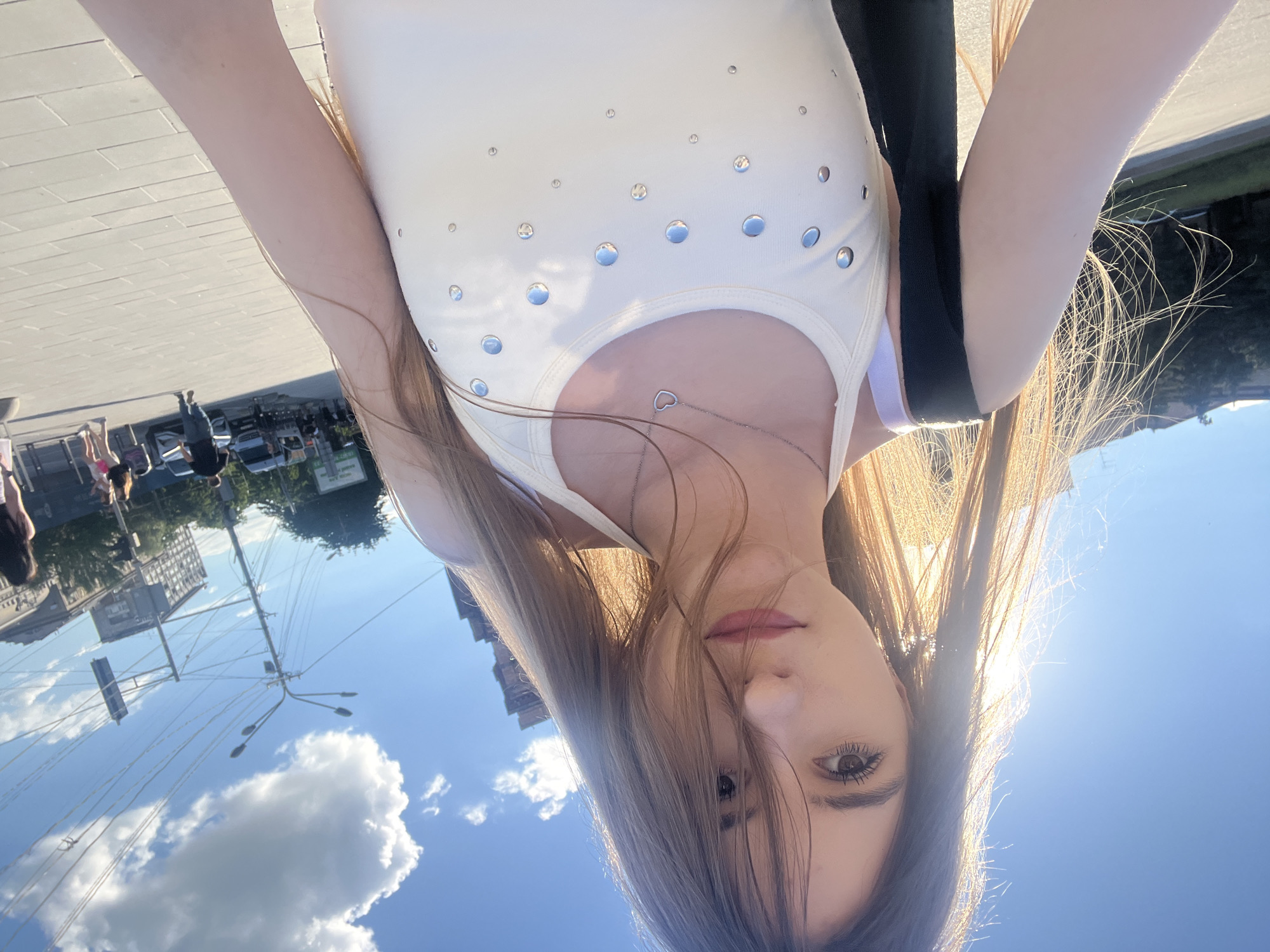 This is a selfie taken outdoors in an urban environment, likely in Dnipro, Ukraine, given the visible street signs with Cyrillic text and architectural style. The sun is bright, casting a strong light that creates a lens flare and highlights the subject's hair.
The subject is a young woman with long, light brown hair, partially obscuring her face. She is wearing a white tank top adorned with silver studs of various sizes. A thin silver necklace with a heart pendant is visible. A black bag with purple straps is slung over her shoulder.
In the background, a paved walkway is visible, with buildings and streetlights. Several other people are walking in the distance, and a few vehicles are present on a street behind them. The sky is blue with some fluffy white clouds. The overall impression is that of a casual daytime scene in a city.