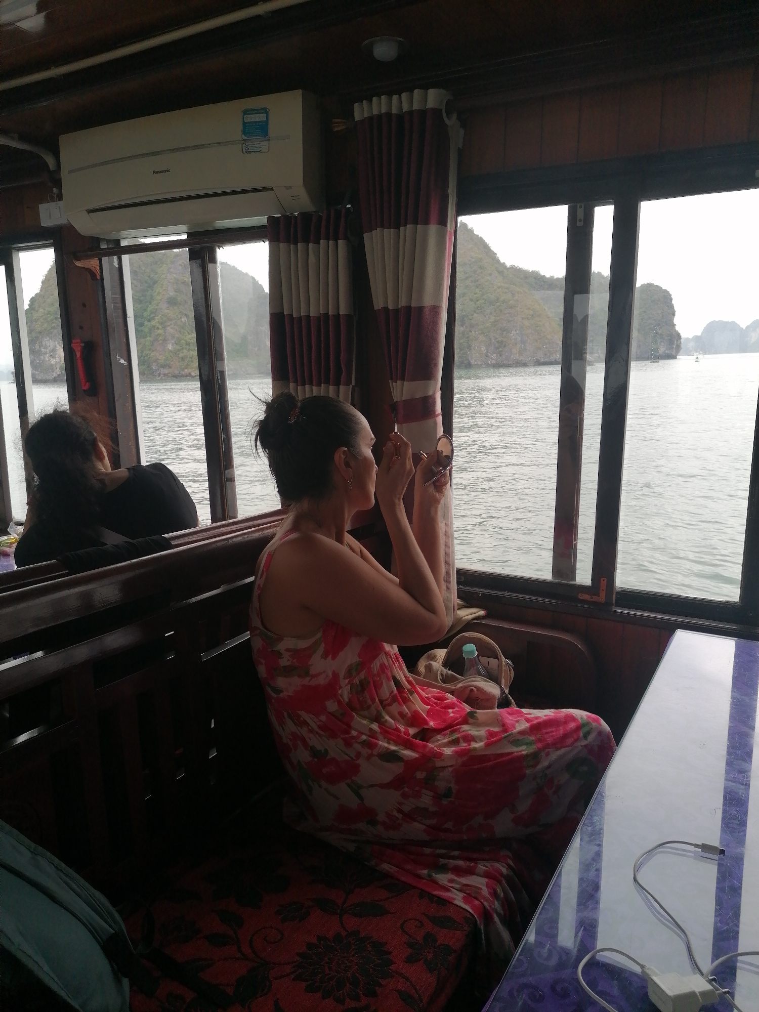 The image captures an interior view of what appears to be a boat or cruise ship cabin, looking out onto a scenic body of water with distinctive limestone islands.
In the foreground on the right, a woman with dark hair tied back in a bun, wearing a sleeveless red and white floral dress, is seated on a wooden bench. She is holding a compact mirror in her left hand and a brush or applicator in her right, actively applying makeup. Beside her on the bench, a light-colored bag containing a water bottle is visible. Behind her, to the left, another woman with dark hair is partially visible, seated and looking out the window, with her back mostly to the camera.
The cabin features a wooden ceiling and window frames, creating a warm, enclosed atmosphere. Red and white striped curtains hang beside the windows. Above the windows on the left, a white Panasonic air conditioner is mounted, displaying a blue label with some unreadable text. The wooden bench is covered with a dark red cushion featuring black floral patterns. On the right, a translucent table, possibly glass, with purple patterns underneath, holds a white charging cable and adapter.
Through the large windows, the calm, grey-blue water stretches out, dotted with characteristic lush green limestone karsts or islands. The sky appears overcast, suggesting a cloudy day, yet it is clearly daytime. The scene is consistent with the landscape of Ha Long Bay, fitting the provided location context of Bai Chay Ward, Vietnam.