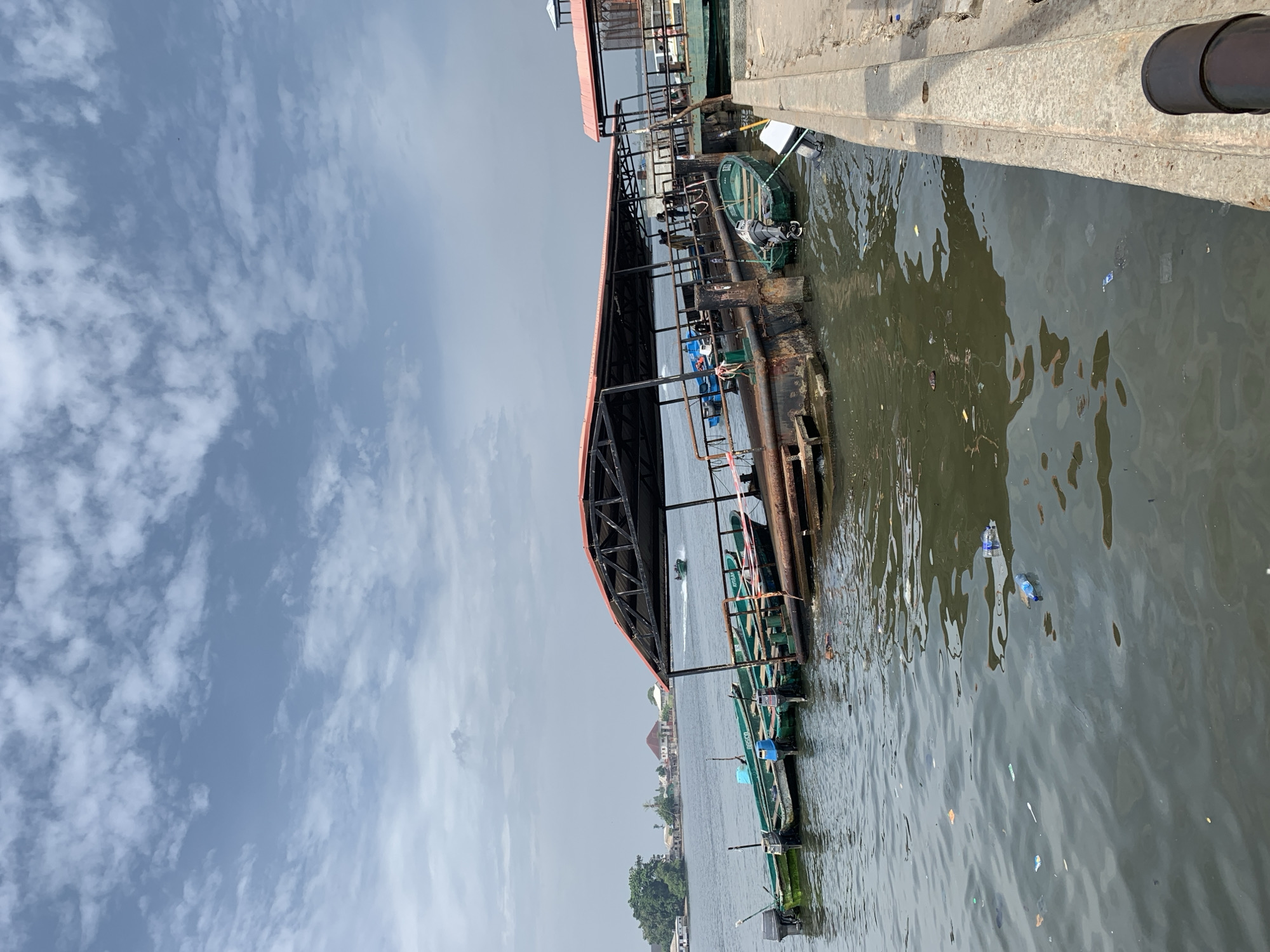 This image depicts a waterside scene at a jetty or pier in Ibasa, Nigeria, under a partly cloudy sky. The main subjects are a long, covered walkway/jetty extending into the water, several small boats, and the surrounding water and distant land.
The jetty, constructed from metal framework and topped with a reddish-brown roof, runs parallel to a concrete or metal pier on the right side of the frame. Several small, mostly green, wooden boats are moored along the jetty. Many of these boats are equipped with outboard motors, suggesting their use for transport or fishing. A few individuals are visible on the jetty, possibly waiting or attending to the boats.
The body of water appears murky and exhibits some surface turbulence. Notably, there is visible debris, including plastic bottles and other trash, floating in the water near the pier and jetty. In the distance, across the water, parts of a landmass with residential buildings and lush green vegetation can be seen, hinting at a coastal or riverine community. One small boat is actively moving in the mid-distance, leaving a faint wake behind it.
The weather is bright and daytime, with a significant portion of the sky covered by scattered, wispy white clouds against a pale blue background. On one of the green boats, the word "IBARAP" is visible on its side, and another boat further down appears to have "IBERECU" or similar lettering. The scene captures a typical bustling waterside environment, highlighting both functionality and environmental concerns.