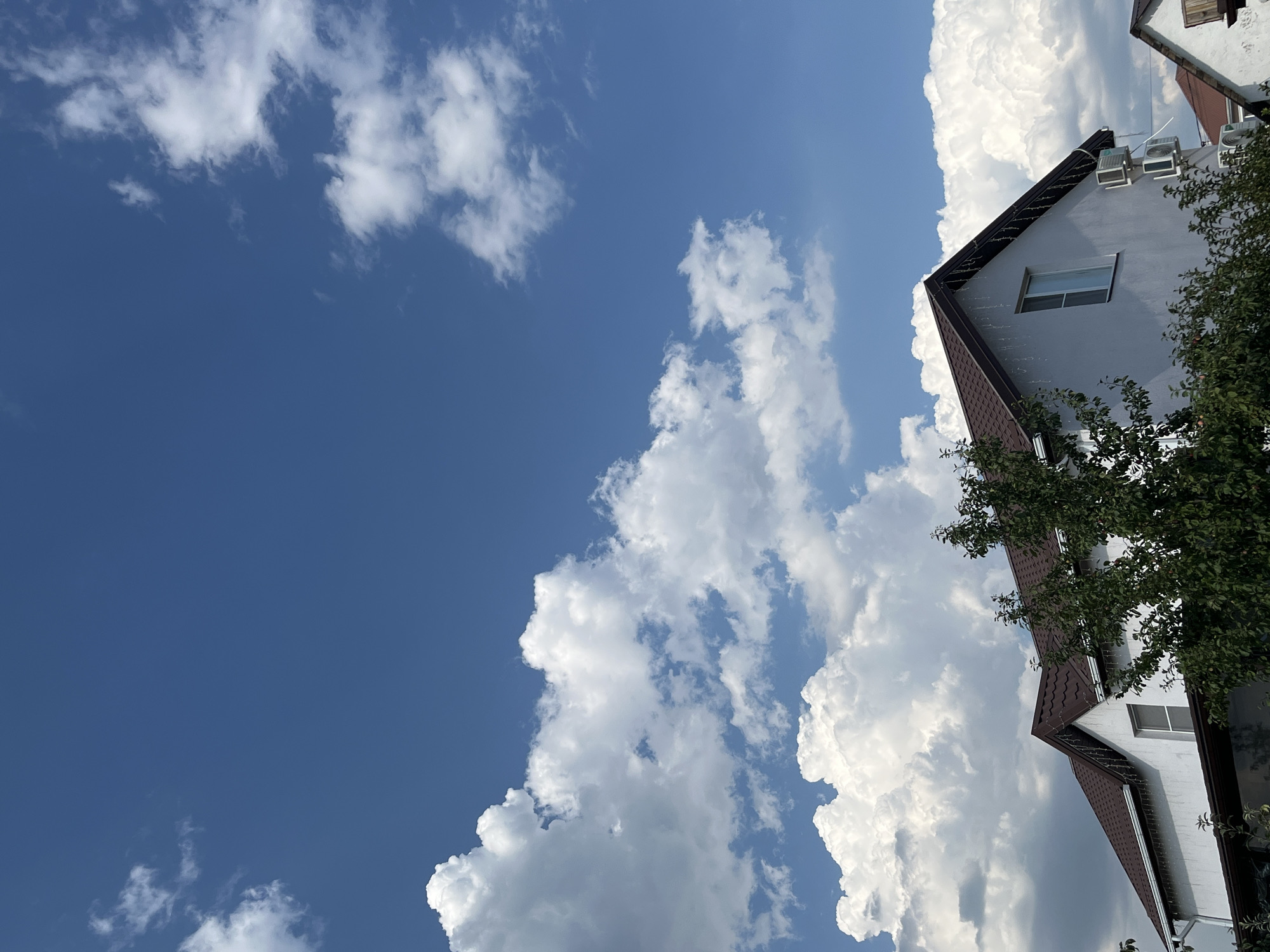 The image depicts a bright, sunny day with a clear blue sky filled with large, fluffy white clouds. The lower portion of the image shows the rooftops of houses, a prominent white building with a brown roof, and several trees with green foliage. There are visible architectural features such as windows, an air conditioning unit mounted on the wall, and gutters. The scene suggests a residential area, likely a suburb or a town, under fair weather conditions. The setting is the city of Taganrog City District, Russia.
