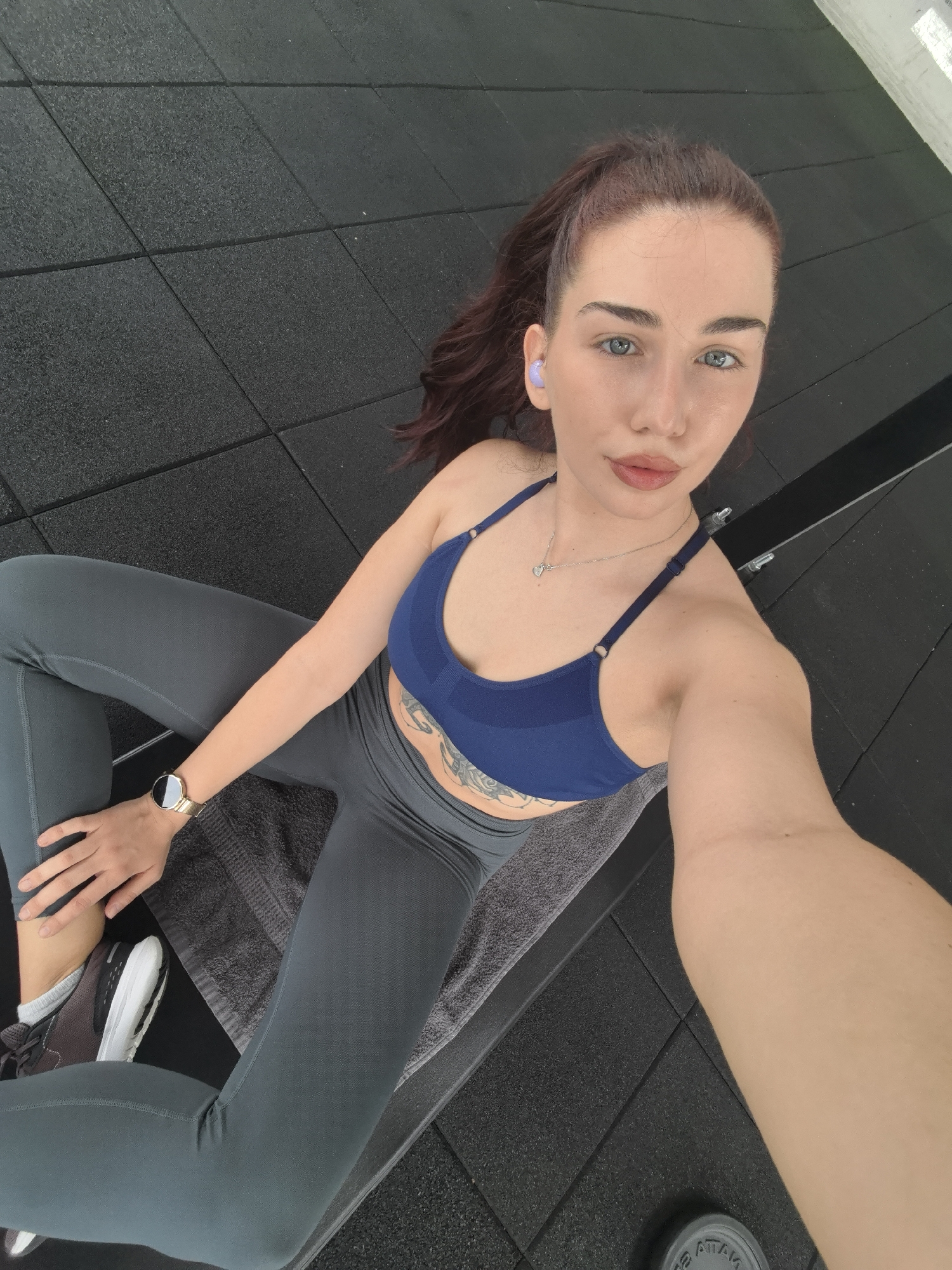 A woman is sitting on the floor of a gym. The floor is covered in black rubber tiles. She is wearing a blue sports bra and gray leggings. She has brown hair pulled back in a ponytail and is wearing earbuds. She has a tattoo on her stomach. She is also wearing a watch. Her shoes are sneakers. A gray towel is between the woman and the black gym floor. A weight is barely visible at the bottom of the frame. The scene appears to be in a gymnasium in Stara Zagora, Bulgaria, though this cannot be confirmed with available details.