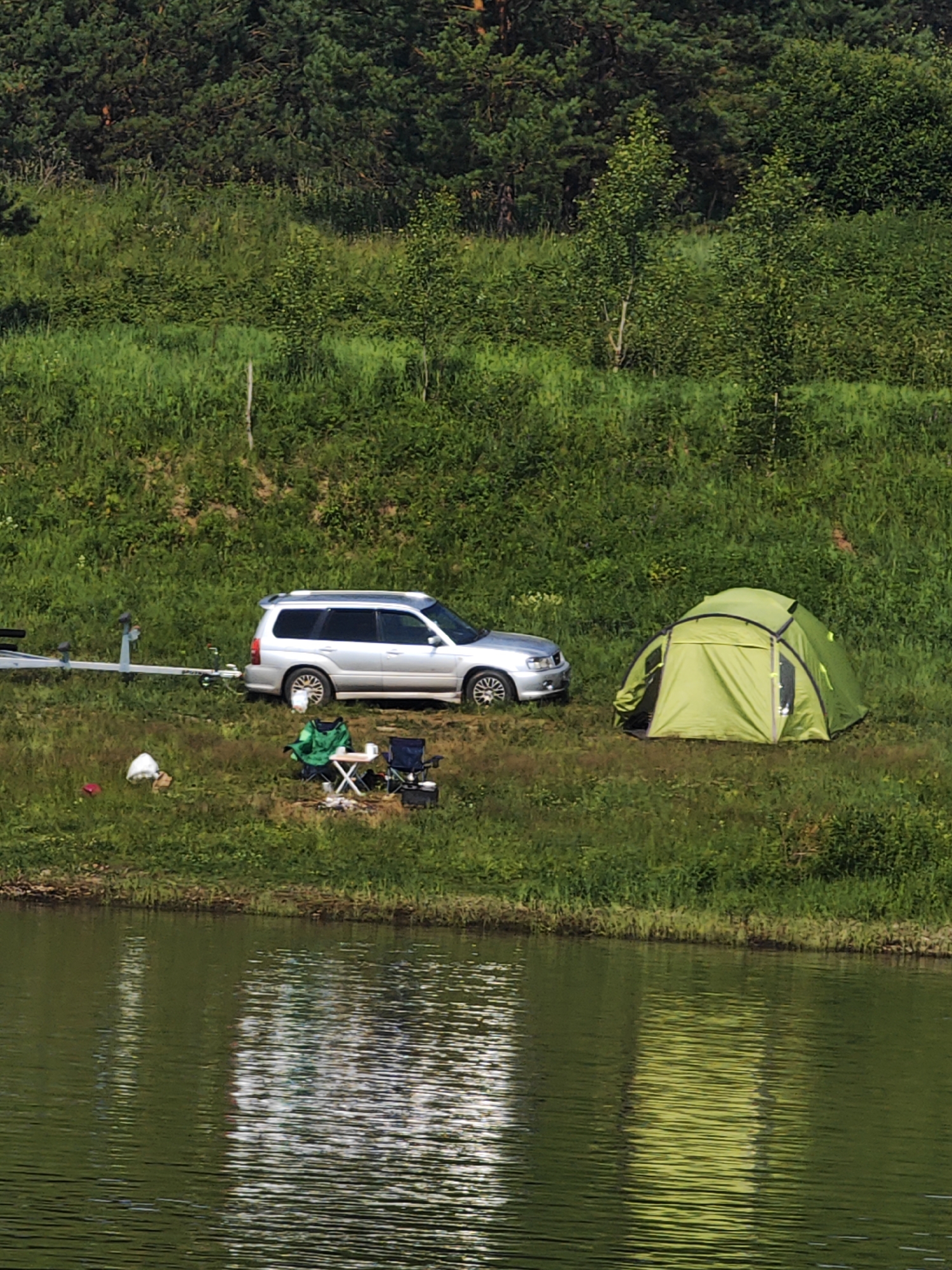 The image depicts a campsite on the grassy bank of a body of water, likely a river or lake, with a dense forest in the background. A silver SUV is parked on the grass, hitched to a boat trailer. A green tent is pitched nearby. In front of the SUV, a person is seated in a green jacket at a small folding table with two chairs, suggesting a moment of rest or activity. Several white plastic bags and a red object are scattered on the grass. The water in the foreground reflects the trees and sky, creating a textured surface. The overall scene suggests an outdoor recreational activity.