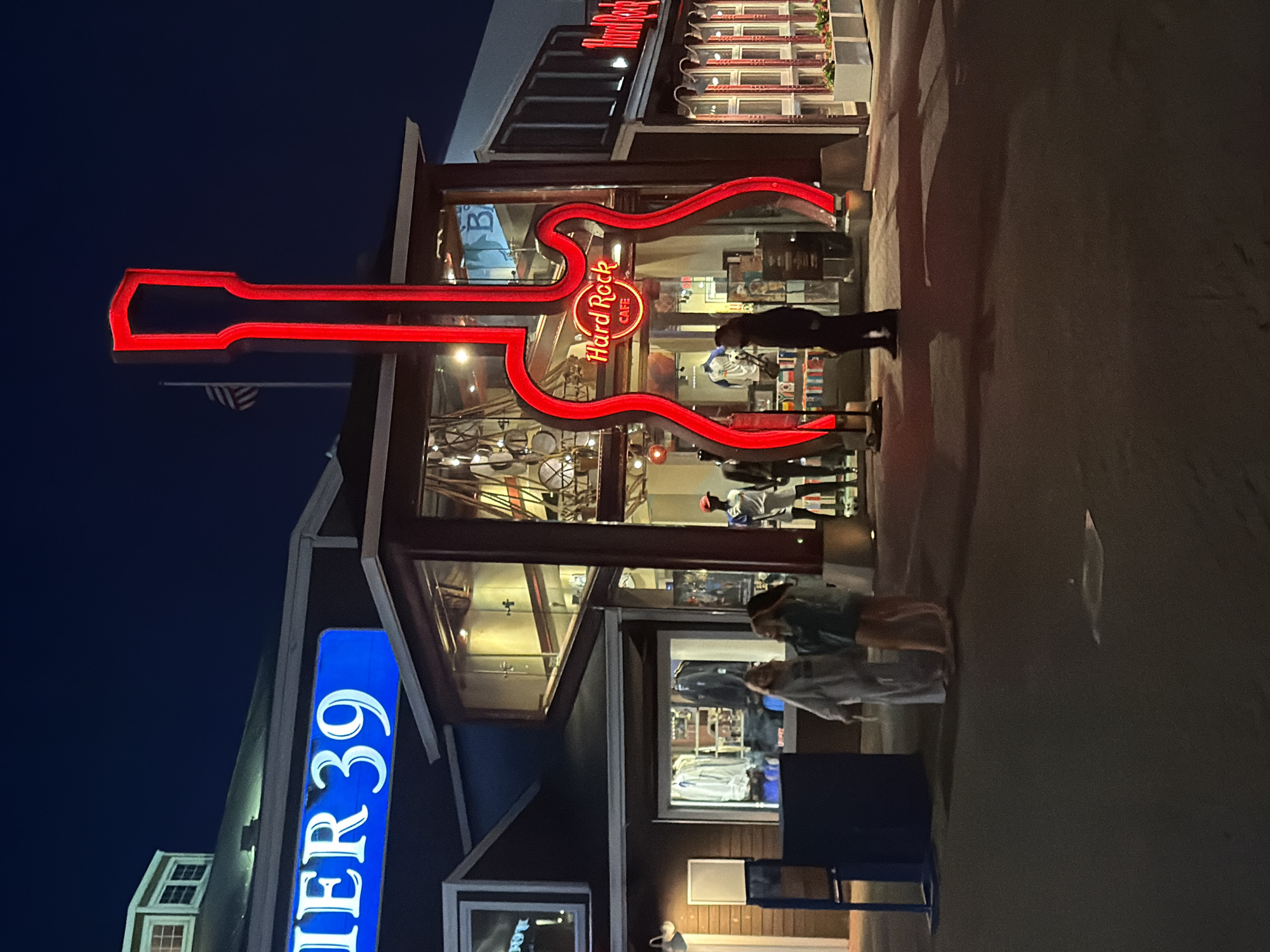 The image depicts the Hard Rock Cafe at Pier 39 in San Francisco, California, USA, at dusk. The iconic red neon guitar sign of the Hard Rock Cafe is prominently displayed in front of the establishment. The building features large glass windows, offering a glimpse into the cafe's interior, which appears to be decorated with various memorabilia. Adjacent to the Hard Rock Cafe, a "PIER 39" sign is visible, illuminated in blue. Several individuals are present outside the establishments, with some appearing to be browsing merchandise displayed in a shop window. The ground in front of the buildings is paved, and a few streetlights cast a warm glow on the scene. An American flag is visible flying on a flagpole in the background. The overall atmosphere suggests a lively evening at a popular tourist destination.