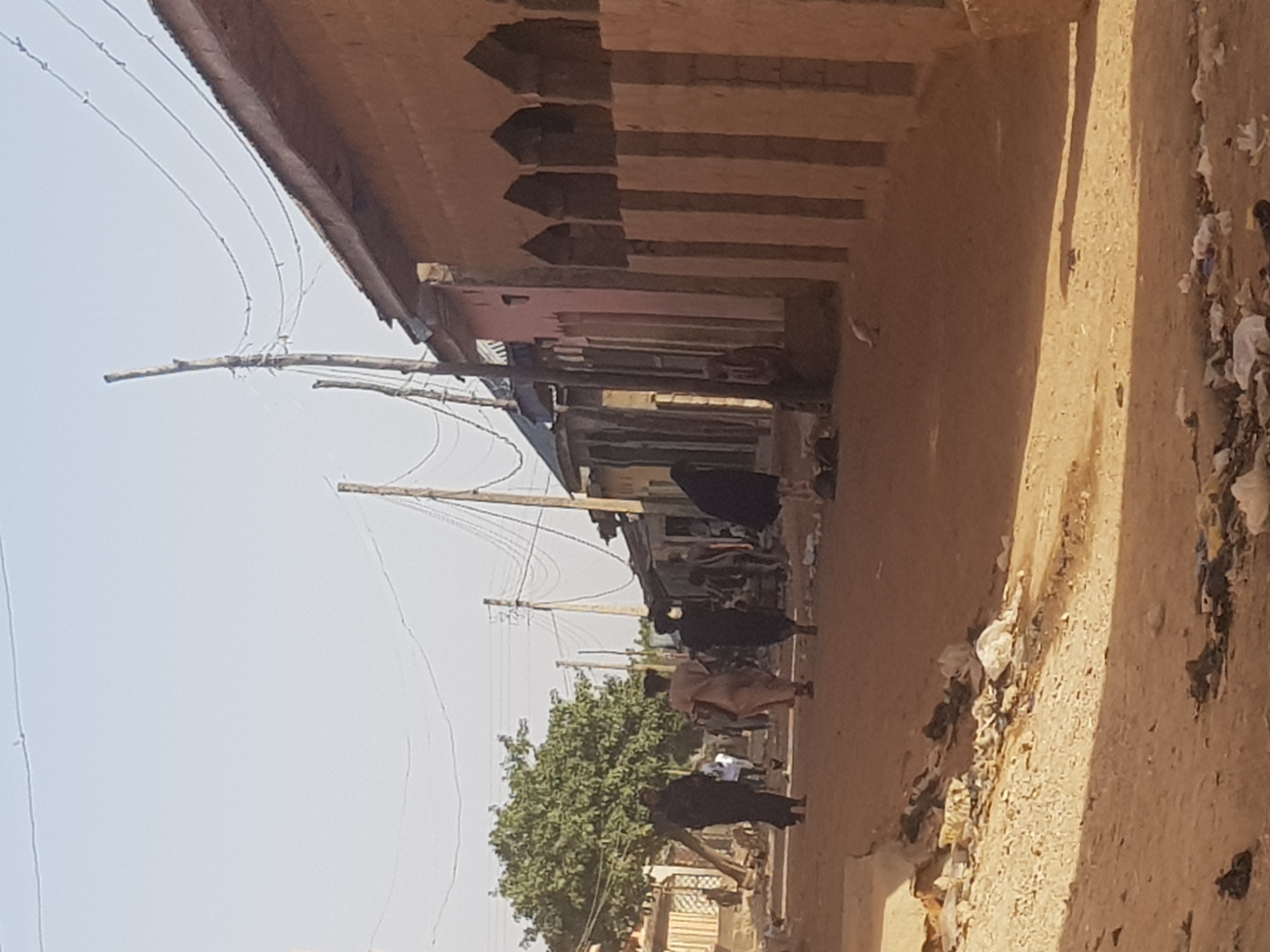 The image depicts a street scene in Dutsinma B, Nigeria. Several individuals, some wearing dark clothing, are walking along a dusty street. A building with a distinctive roofline is visible on the left. Power lines run across the sky. The street is littered with debris.