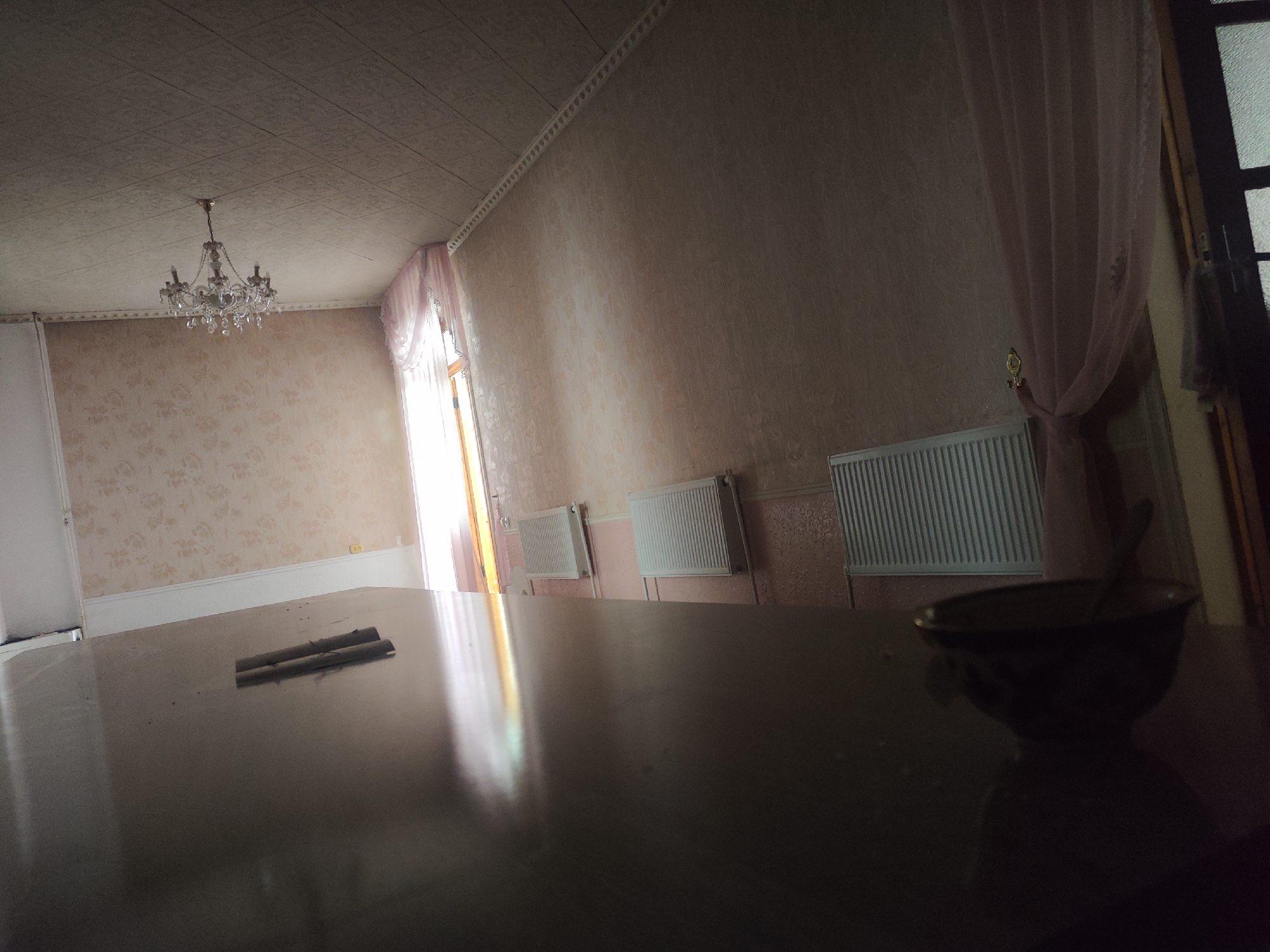 A dimly lit room with a chandelier hangs from the ceiling. The walls are covered in patterned wallpaper, and there are radiators along one wall. A large dark table dominates the foreground, reflecting the light from a window on the left. On the table, two cylindrical objects are placed side-by-side, and a dark bowl with a spoon rests to the right. A sheer pink curtain is visible on the right side of the room, near a dark doorframe. The scene appears to be an interior of a room in Shahimardanabad, Uzbekistan.