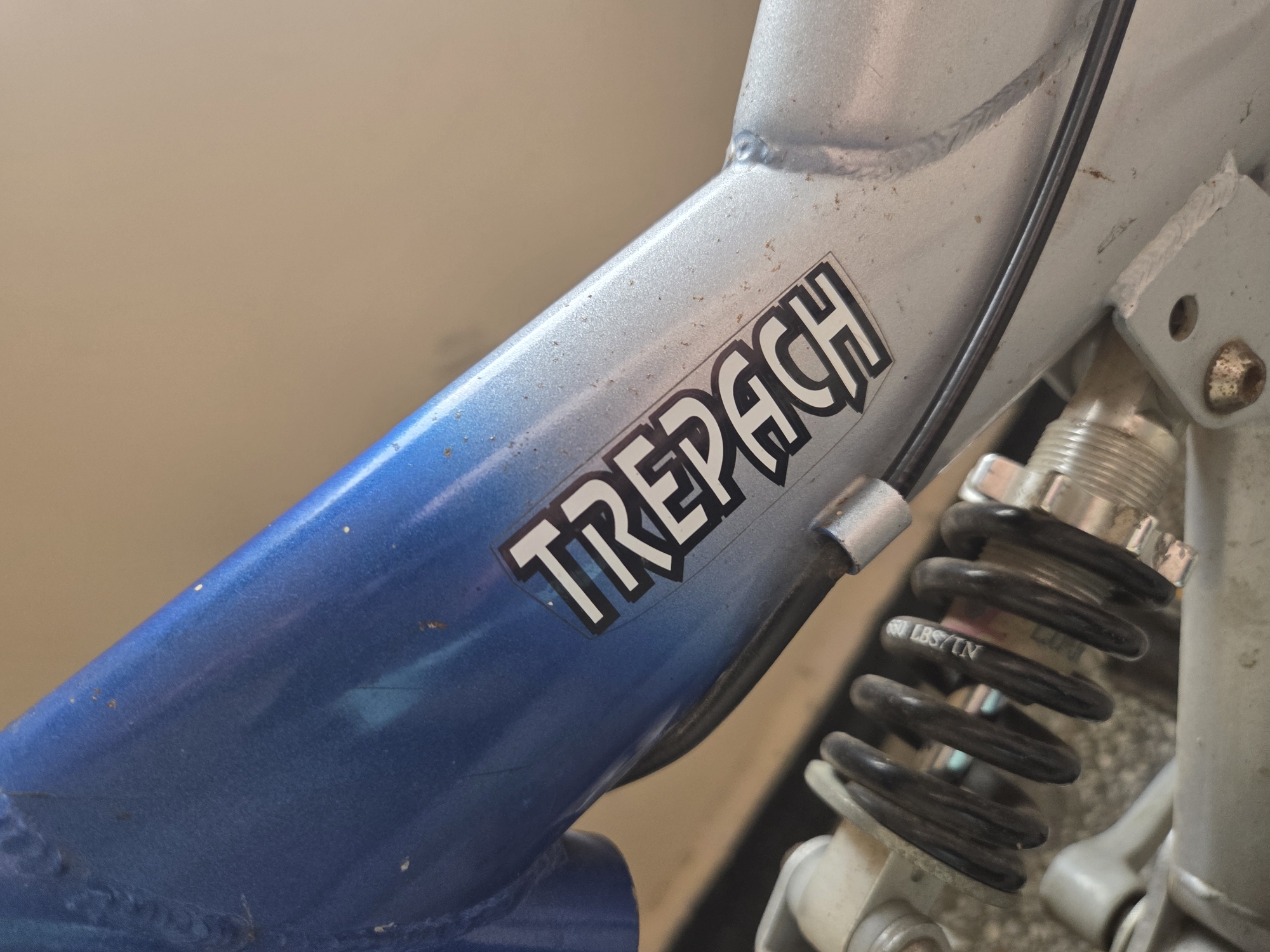 This close-up image captures a section of a bicycle frame, featuring a distinctive paint fade from a vibrant blue at the bottom left to a metallic silver towards the top right. The frame shows signs of wear, including minor scratches, paint chips, and what appear to be small rust specks on the silver areas.
A prominent black sticker with bold, white, stylized text is affixed to the frame, clearly displaying the word "TREPACH". To the right of the frame, a black coil spring, part of a suspension system (likely a rear shock), is visible. White text printed on this spring indicates its rating as "650 LBS/IN". A black cable runs along the silver section of the frame, held in place by a small silver clip. Visible welds on the frame joints highlight its construction.
The background is a softly blurred, light brown or beige surface, suggesting an indoor or sheltered outdoor setting. No people are present, and no specific activity is taking place; the image focuses purely on the bicycle components. The lighting appears to be ambient, possibly natural daylight. The photograph was taken in Pleven, Bulgaria.