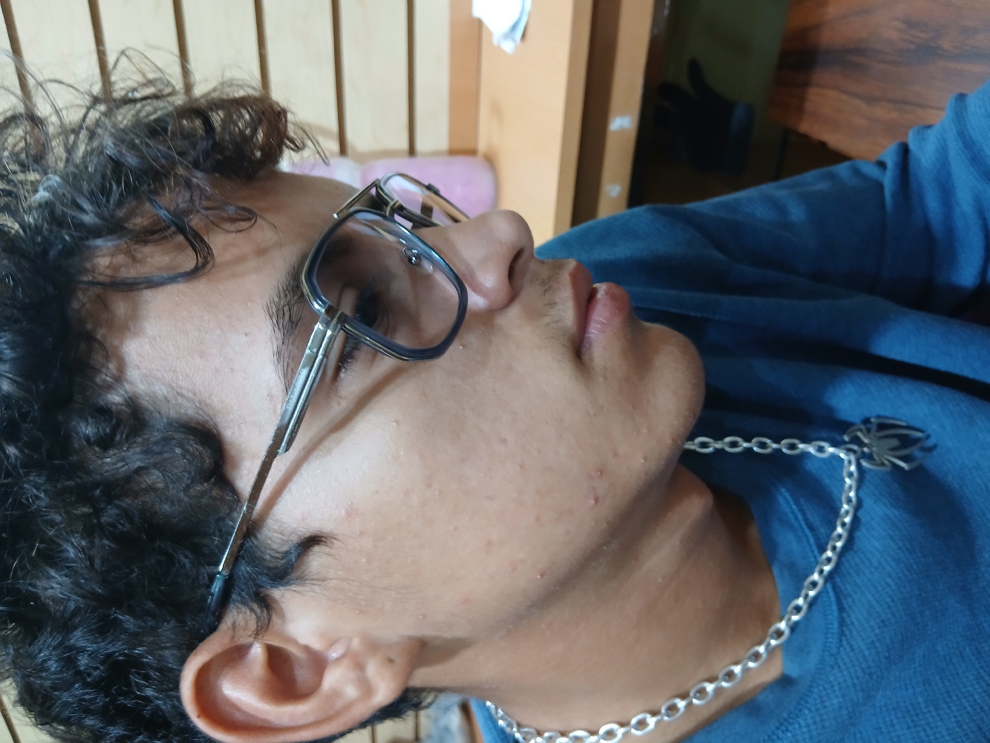 The image shows a close-up, side profile of a person wearing glasses and a blue shirt. The person has dark, curly hair and appears to be looking to the side, with their mouth slightly open. A silver chain with a spider pendant is visible around their neck. The background suggests an indoor setting with wooden elements. The lighting indicates it could be daytime, and the overall mood is relaxed. There is no visible text in the image. The person's skin has some minor blemishes.