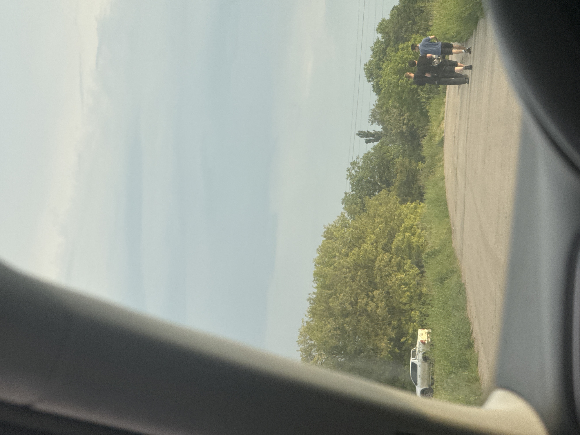 The image captures an outdoor scene in Suprunivka, Ukraine, from the interior perspective of a vehicle, indicated by the dark car dashboard and A-pillar visible in the foreground. The view is directed along a two-lane, unpaved or partially asphalted road bordered by tall green grass and trees with light green foliage.
On the left side of the road, off the main path and situated within the tall grass, a white four-door pickup truck is parked and stationary. Further down the road, walking away from the camera, are three male individuals. The leftmost person is wearing a dark shirt and dark pants. The middle individual is attired in a dark shirt and dark shorts, with a dark bag or pouch visible at their waist. The rightmost person is wearing a blue shirt and dark shorts. All three are walking in the same direction, along the road.
The background consists of dense green tree lines under a light blue, partly cloudy sky. Utility poles and power lines are visible above the trees on the right side of the frame. The lighting suggests daytime conditions.