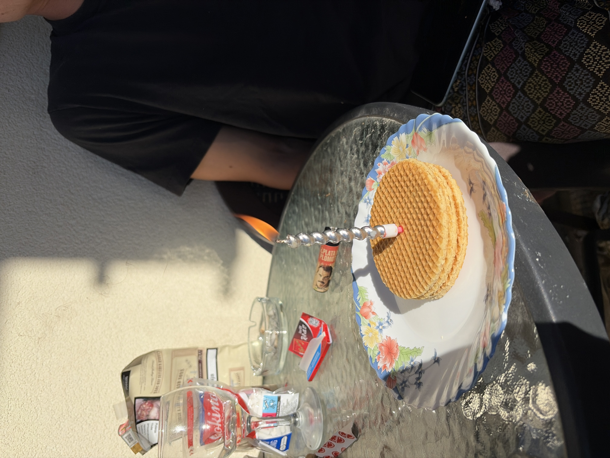 The image captures an outdoor scene in Budva, Montenegro, under bright daylight with direct sunlight casting distinct shadows. A round outdoor table with a glass top occupies the central and foreground areas. On the table, a white ceramic plate with a blue and floral rim holds a stack of four round, grid-patterned wafers. A lit, silver, spiral-shaped birthday candle with a red base is inserted into the uppermost wafer. To the left of the plate, a clear wine glass, a plastic bag of rolling tobacco with visible health warnings and text "GEORGE KARELIAS AND SONS," an empty glass ashtray containing multiple cigarette butts, a small open red booklet of rolling papers, and a small, dark cigar stub with a label depicting a face and the text "PLUM" are arranged. A person is partially visible on the right side of the frame, seated at the table, wearing a black short-sleeved t-shirt. The lower half of their visible body shows patterned pants with a geometric design. A smartphone is partially visible on their lap or beside them. The background features a textured, light-colored wall, upon which sharp shadows from an unseen vertical structure are cast.