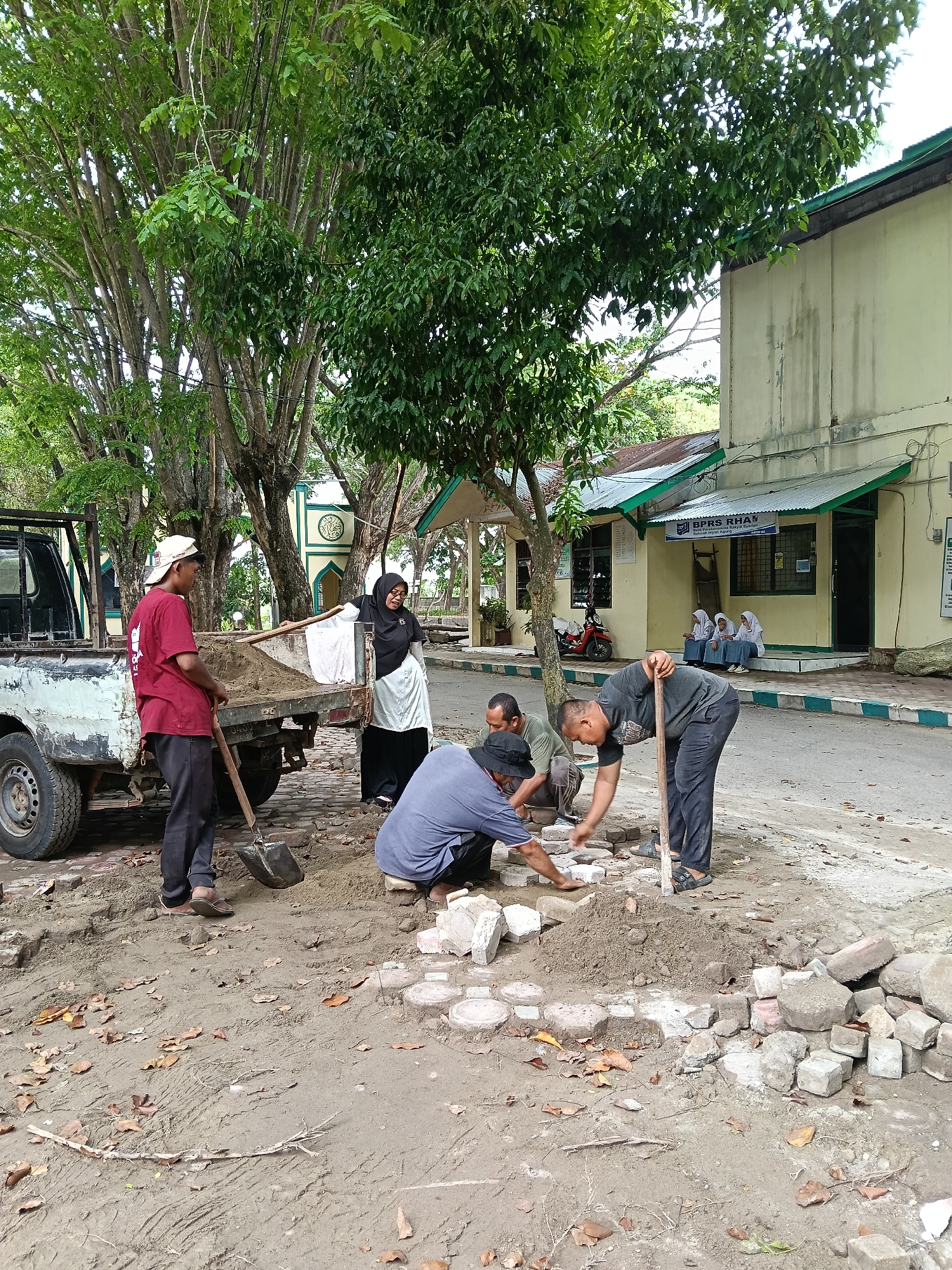 In Lhokseumawe, Indonesia, a group of five adults are working on a construction or paving project in an outdoor area near a building and trees. One person, wearing a red shirt and light-colored cap, stands near a small, white pickup truck loaded with what appears to be sand or gravel. He holds a shovel. Nearby, a woman in a black headscarf (hijab) and long black skirt stands next to the truck. Three other men are actively engaged in laying or adjusting paving stones or bricks on the ground. One man is kneeling, another bent over, and a third is in the background. A pile of stones and bricks sits nearby. In the background, students in uniforms are sitting on the steps of a building with a sign that reads "BPRS RHA." A mosque can be seen partially obscured by trees. The ground is uneven, covered in dirt and scattered leaves.