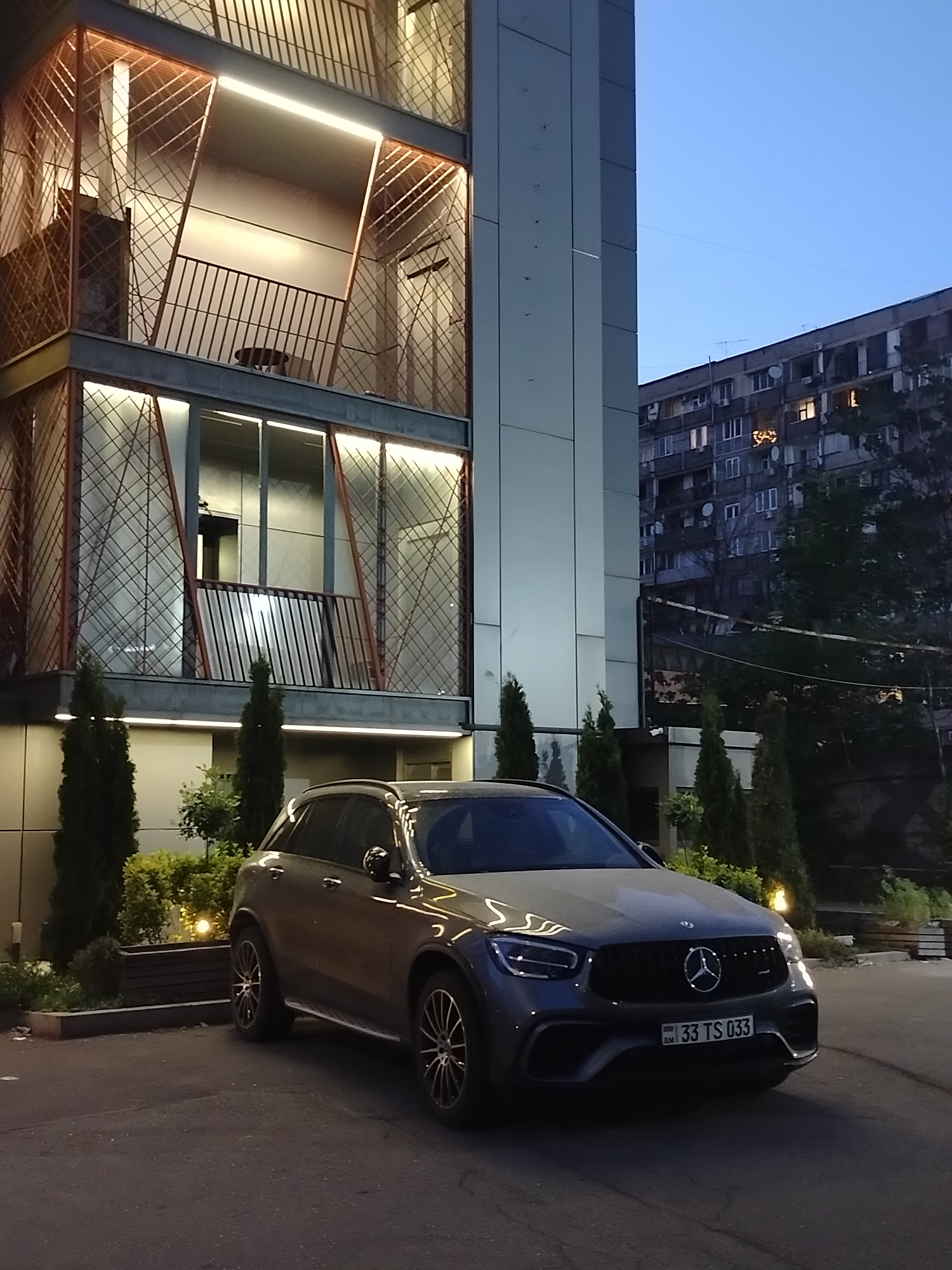The image depicts a modern architectural building with a dark gray Mercedes-Benz GLC parked in front. The building features a distinctive facade with geometric patterns and illuminated balconies. A multi-story apartment building with visible windows and a partial view of trees is in the background. The scene is set in Yerevan, Armenia, as indicated by the Armenian license plate "33 TS 033" on the car. The light suggests it is either dusk or early evening. No people are visible in the image.