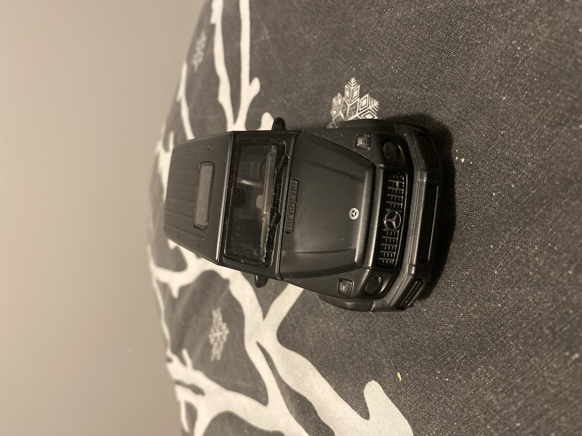 A black Mercedes-Benz G-Wagon toy car is positioned on a patterned cloth surface. The cloth has a dark grey base with abstract white markings resembling branches and snowflakes. The toy car is viewed from a slightly elevated, front-facing angle, showcasing its grille, headlights, windshield, and roof. The background is a plain, light-colored wall. There is no indication of the city of Łódź, Poland, or any people present in the image.