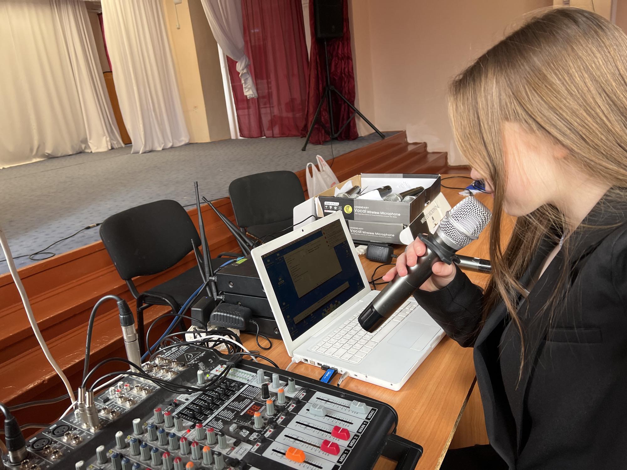 The image depicts a person, likely a young woman, holding a wireless microphone near a laptop computer and an audio mixing console. The scene appears to be a setup for sound or audio control, possibly in a venue or stage environment.
Here are the details:
People:
- A young woman is visible on the right side of the image. She is holding a microphone.
Subjects:
- The main subjects are related to audio and sound equipment.
Objects:
- Wireless microphone (held by the woman and in a box)
- Laptop computer (white)
- Audio mixing console
- Boxes of wireless microphones
- Cables and wires
- Chairs
- Stage-like backdrop with curtains
- Speaker on a stand in the background
Actions:
- The woman is holding a microphone and may be testing or adjusting sound settings.
- Interaction with equipment: Likely adjusting or monitoring the sound system.
Environment:
- The background suggests a venue, hall, or stage setup. There are stairs leading to the stage.
- The location is Chaikovsky, Russia (implied by the image content).