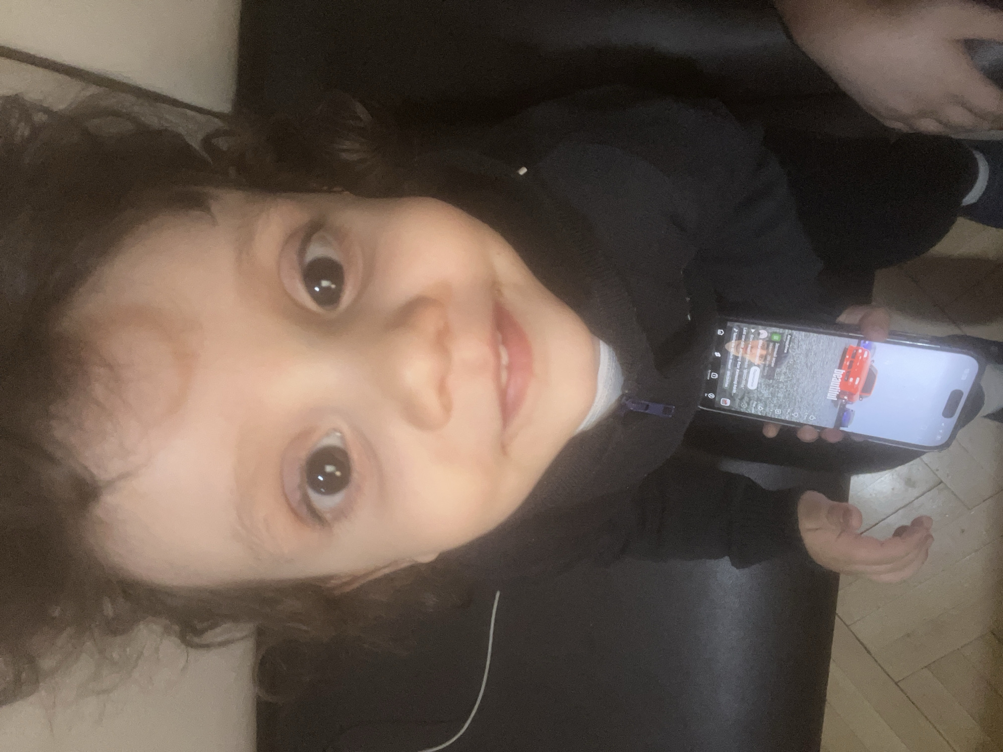 A young child with curly dark hair and brown eyes is holding a smartphone in their right hand. The phone screen displays a video game with a red car. The child is wearing a black hooded sweatshirt. The child is sitting on a black couch. Another person's arm and hand are visible in the upper right corner of the image, possibly belonging to an adult. The flooring appears to be wooden. Location is Unknown, Unknown.