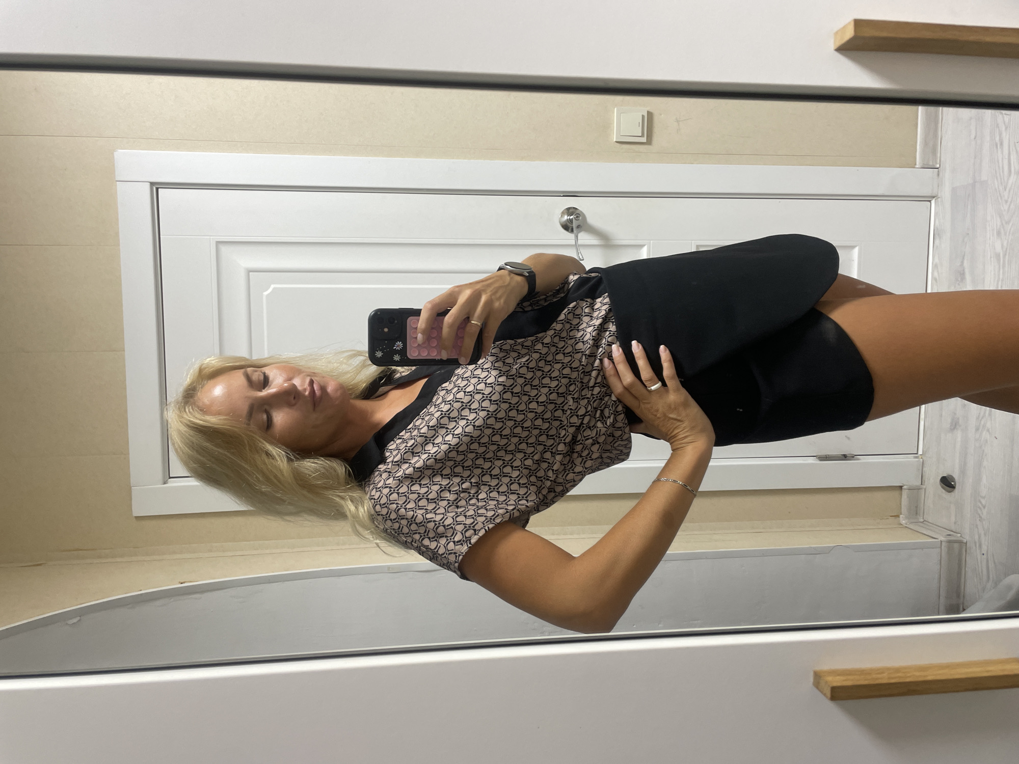 In a domestic interior environment, a blonde woman with fair skin stands before a mirror, taking a selfie. She is wearing a black wrap skirt and a short-sleeved blouse with a geometric pattern in beige and black. The woman has her left hand on her hip and her right hand holds a smartphone. The background features a white door, beige walls, and a white baseboard. The flooring appears to be light-colored wood. This scene does not provide any information that can be used to determine a specific location within Baranovka, Russia.
