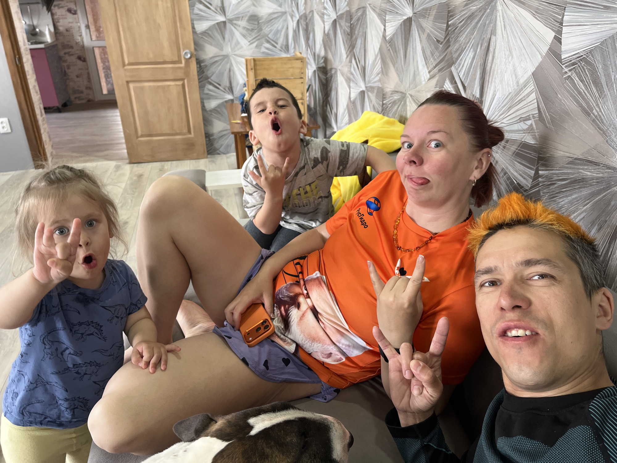 This media file is a lively indoor self-portrait featuring four people and a dog, set in what appears to be a living room or common area of a home.
The subjects include two adults and two children, all striking playful poses for the camera. On the left, a young girl with light hair, dressed in a blue t-shirt with dinosaur prints and yellow shorts, holds up her fingers with a surprised expression. Behind her, a young boy in a camouflage t-shirt makes a "rock on" hand gesture, his mouth wide open in a playful yell.
In the center, a woman with dark hair pulled back, wearing an orange t-shirt, purple shorts with heart prints, and an amber-colored beaded necklace, sticks out her tongue and makes a "rock on" gesture. Her orange t-shirt displays a graphical print that resembles an animal or face, and partially visible text reads "РусМиро" (RusMiro) alongside a circular logo. To the right, a man with short, bright orange-dyed hair and a dark patterned long-sleeved shirt also makes a "rock on" gesture, looking directly at the camera with a slight smile. A dog with white and brown fur is partially visible in the bottom-center, resting its head.
The background features a distinctive grey and white wallpaper with an abstract, geometric pattern resembling sunbursts. A wooden door is visible, and through an open doorway on the left, a glimpse of a kitchen with pink cabinetry can be seen. The overall atmosphere is informal, relaxed, and cheerful, suggesting a fun family moment. The lighting indicates it is daytime.