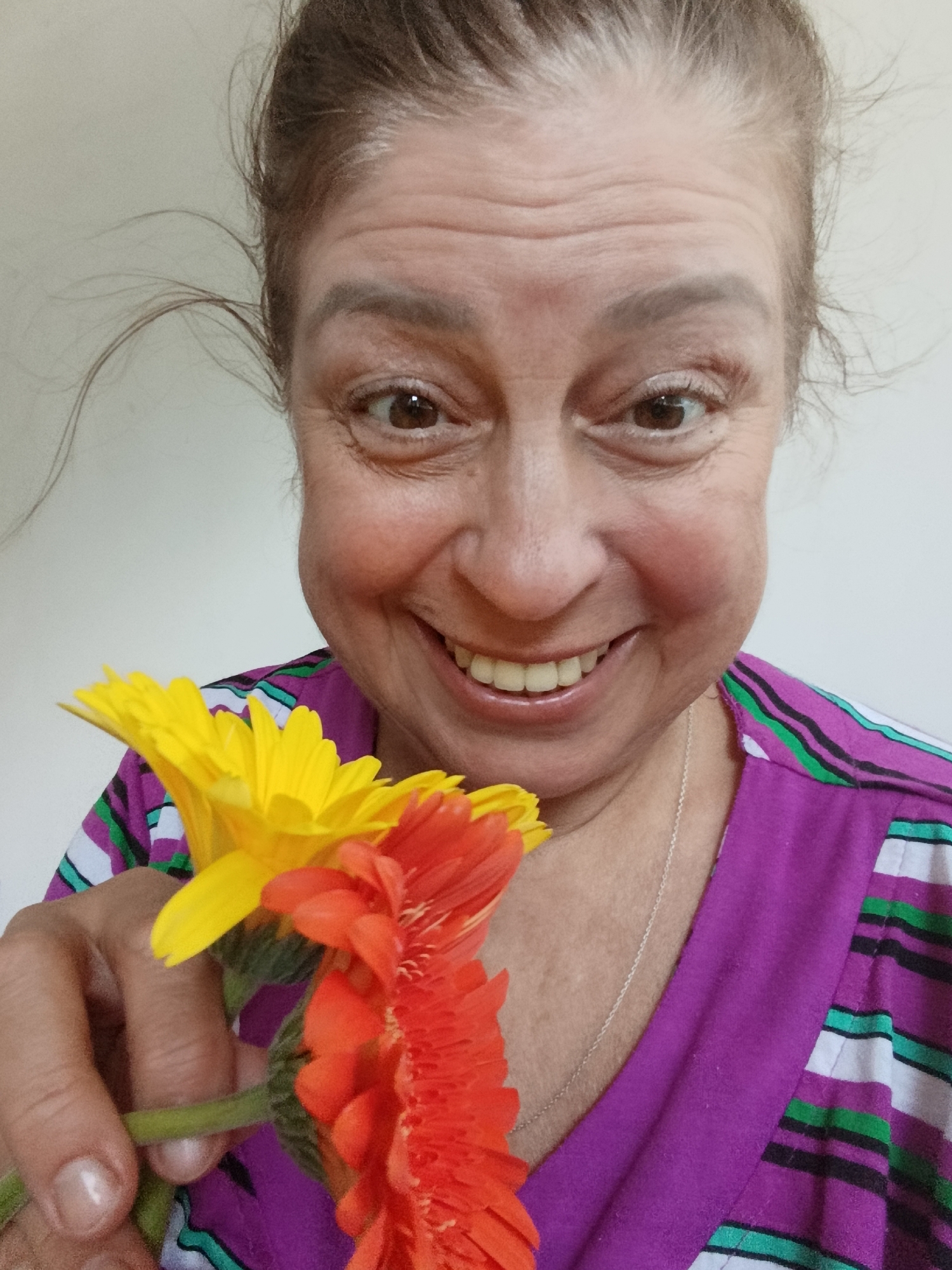 The image shows a close-up, selfie-style shot of a woman holding two gerbera daisies, one yellow and one orange-red, in front of her face. The woman has brown hair pulled back, and she is smiling with her teeth showing. Her eyes are brown and she has wrinkles around them. She is wearing a purple shirt with horizontal stripes of white, black, and green. The background is a plain, light-colored wall. There is no indication of the location being Saratov, Russia.