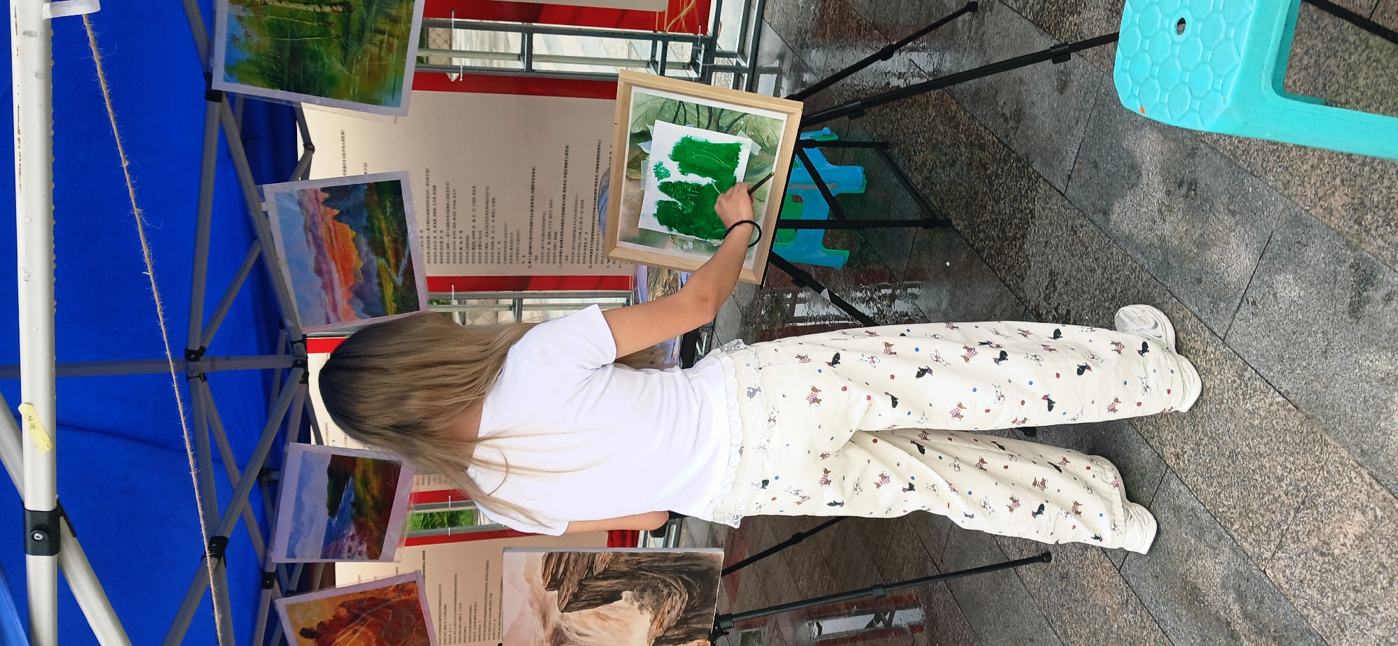A young woman in a white t-shirt and patterned pants stands painting on a framed canvas in an outdoor setting. The canvas is mounted on a black metal easel, and the woman is using a brush to apply green paint. Surrounding her are other framed paintings displayed on what appears to be metal grid stands. A blue plastic stool is visible nearby. The scene takes place on a paved surface, which shows signs of recent moisture, possibly rain. A blue tent or canopy structure is partially visible in the background. The setting seems to be an outdoor art exhibition or display. The location is likely in the Chongqing High-tech Zone, China, based on the query context.