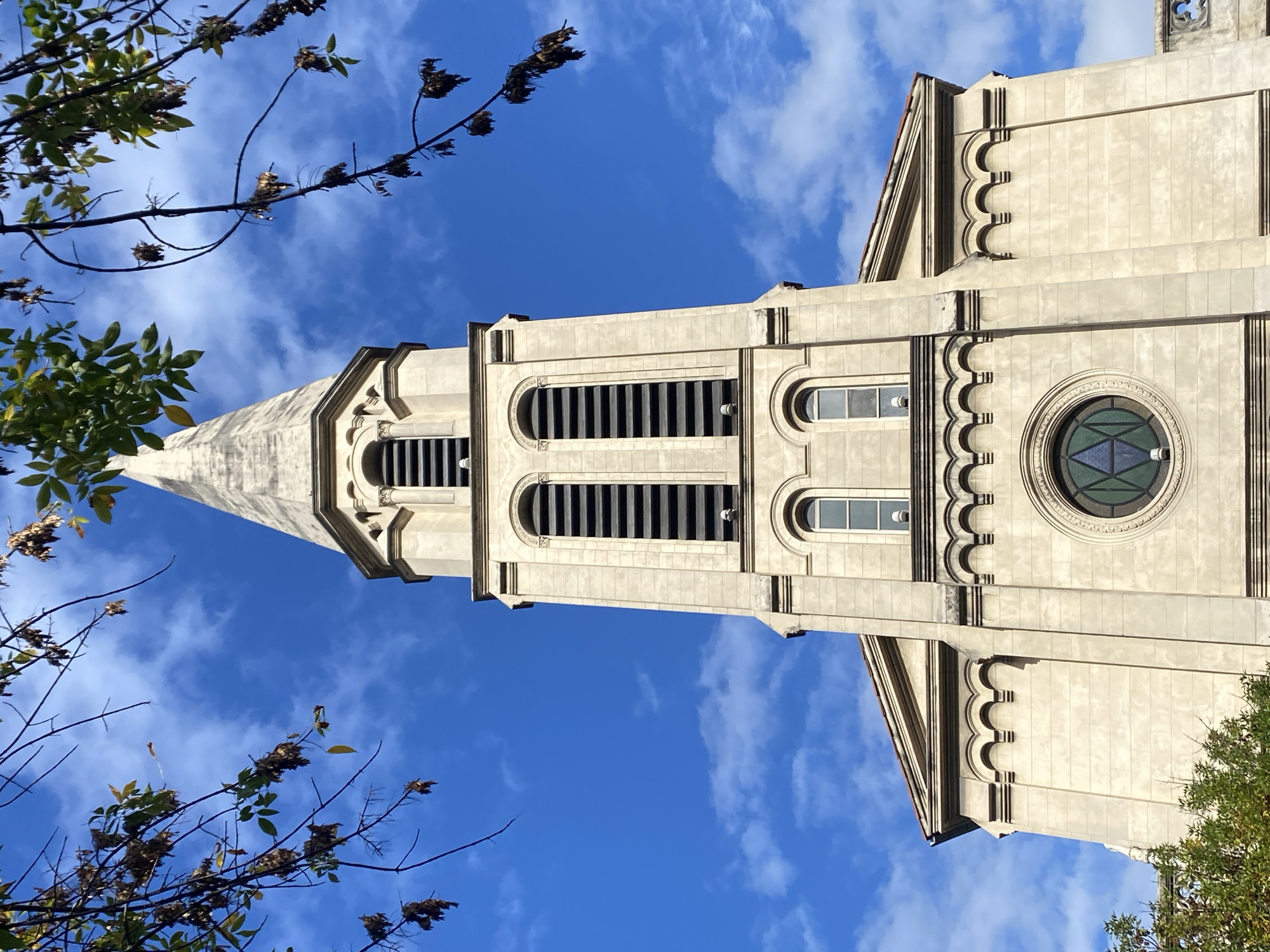A low-angle, full shot presents the First Methodist Church in Buenos Aires, Argentina, set against a backdrop of a clear blue sky with scattered white clouds. The beige church tower, decorated with curved architectural details, rises vertically, interrupted by tree branches with brown and green leaves on both sides of the frame. The tower features rectangular and arched windows, and a round window with stained glass is visible on the lower part. The architecture suggests a gothic or neo-gothic style with a pointed spire at the very top.