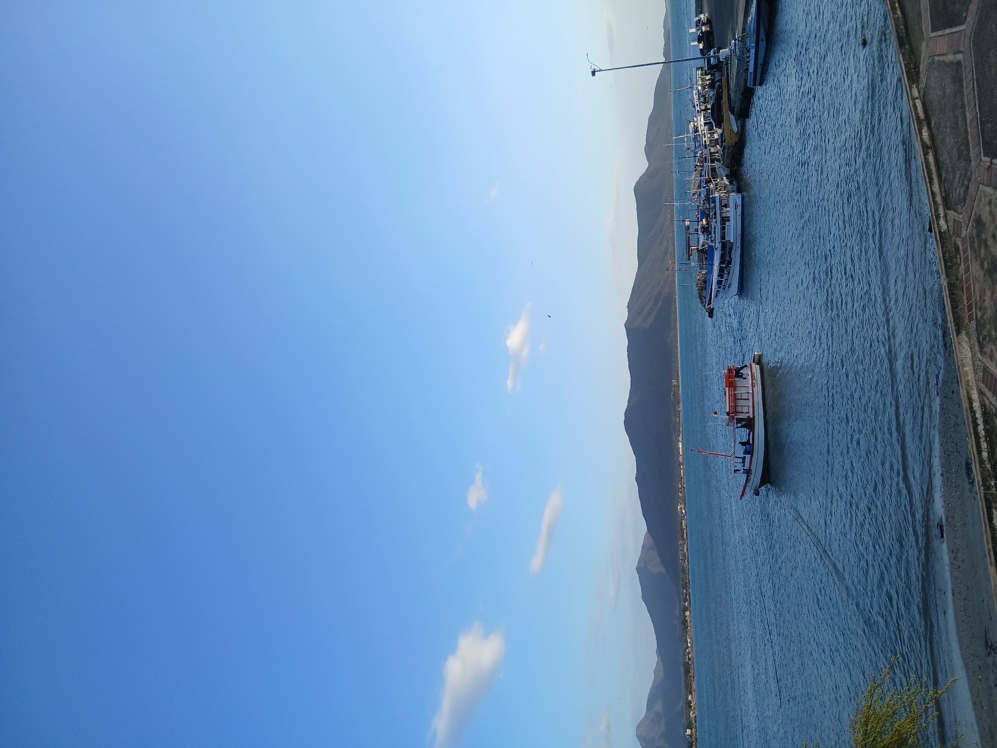 The photograph captures a coastal scene in Juan Griego, Venezuela, presenting a blend of natural landscapes and human elements. Predominantly, the image features a body of water, possibly a bay or inlet, with boats docked along the shoreline. The water's surface is moderately choppy, and the reflection of the sunlight lends it a shimmering appearance.
Several boats are visible, varying in size and style, suggesting a mix of fishing vessels and possibly tourist crafts. On the far side of the water, a mountain range rises, forming a scenic backdrop. Buildings are discernible along the coastline, indicative of urban development integrated with the natural environment.
The sky dominates a significant portion of the frame. The composition is well-lit, highlighting the details of the boats, the water, and the mountainous landscape.