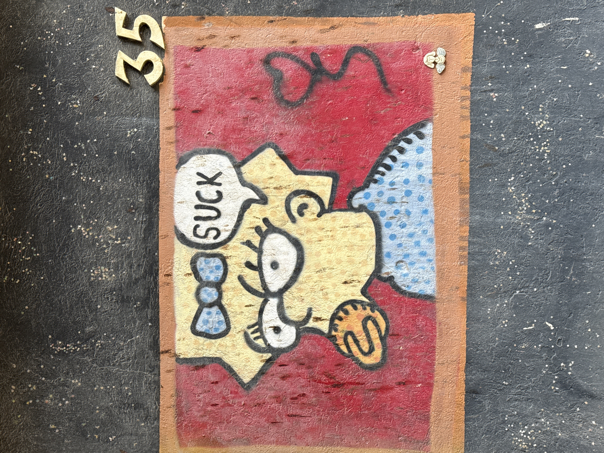 The image shows a crude drawing of Lisa Simpson with a speech bubble saying "SUCK", on a red background with some other scrawled black lines. The drawing is framed with brown. In the top left corner are the numbers "35" in gold. In the upper right corner there is a winged head in gold. The background appears to be a speckled gray surface. Based on the subject matter and style, this image likely represents street art or graffiti. The location is Tel-Aviv, Israel.