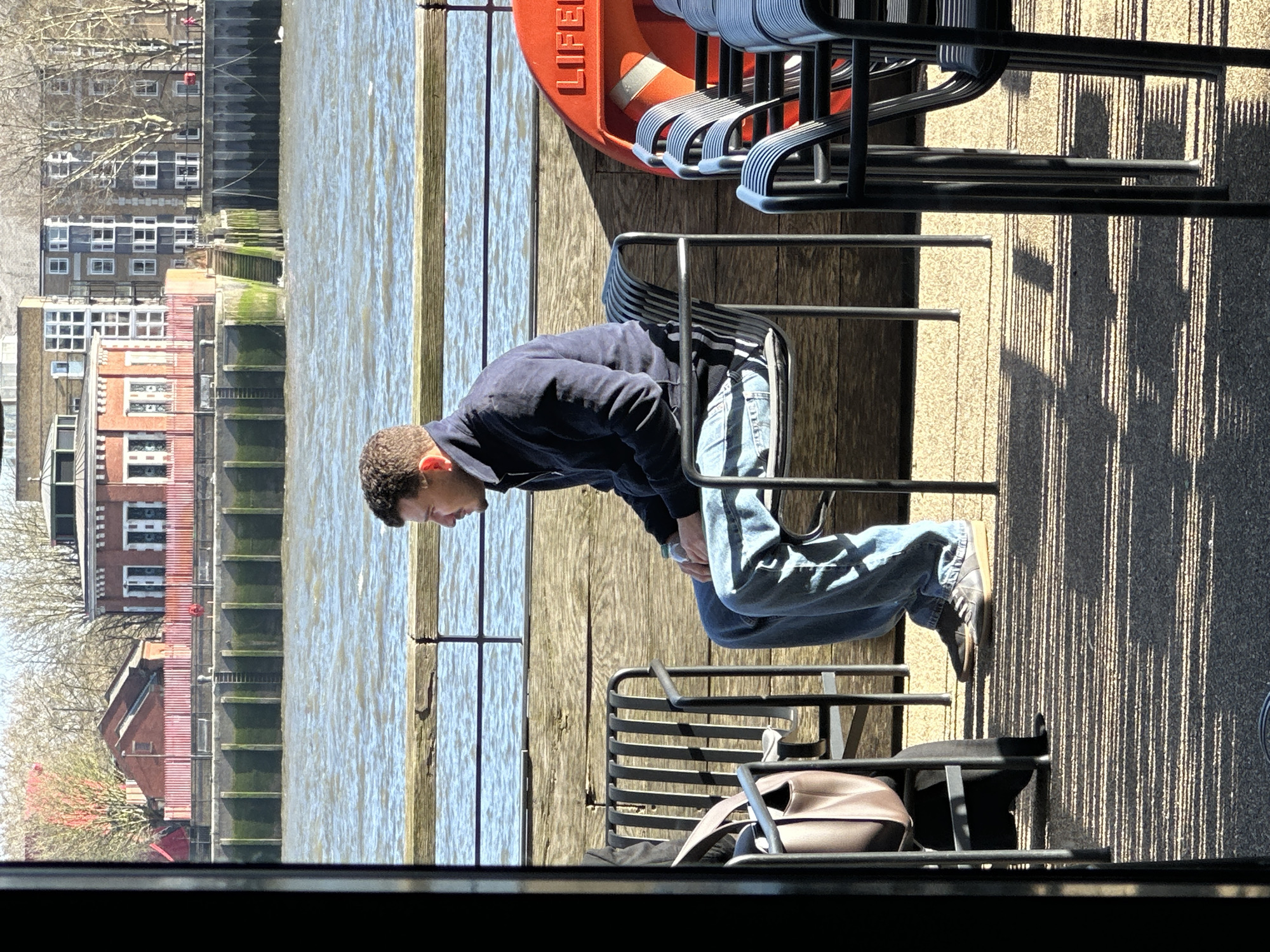 A medium outdoor shot captures a male individual seated in profile on a dark metal chair, facing right, positioned alongside a body of water identified as the River Thames in London, United Kingdom. The man, with light brown curly hair, wears a dark blue zip-up jacket, blue jeans, and dark sneakers. A small, light-colored earbud or earphone is visible in his right ear. His right hand rests on his left thigh, possibly holding a clear plastic bottle. To his left, a dark-clad leg and a brown leather handbag are partially visible, indicating the presence of another individual outside the frame.
A substantial weathered wooden railing separates the immediate seating area from the river. To the right of the seated man, an orange lifebuoy, partially labeled "LIFE", is mounted on a stand. Adjacent to the lifebuoy is a stack of several identical dark metal chairs. The ground beneath is a light-colored, possibly concrete or aggregate surface, displaying prominent parallel shadows cast by the chairs, indicative of direct, low-angle sunlight.
Across the River Thames, the opposite bank features a concrete or stone river wall with vertical wooden pilings, some exhibiting green algal growth. A metal ladder descends into the water on the left side of the far bank, and another set of steps is visible further right. Background structures on the far bank include a red-brick building with a pointed roof, partially obscured by red netting or scaffolding. To its right is a distinctive red-brick building with white trim, arched windows, and a rounded roof section, fronted by a red barrier. Further right, a series of multi-story residential buildings with numerous windows, varying in color (grey, dark blue), are visible. Numerous deciduous trees, currently bare, are scattered across the far bank. A small white object, consistent with a swan, floats on the river to the far right. The scene is partially framed on the left by a dark vertical structural element, suggesting a view through a doorway or window.