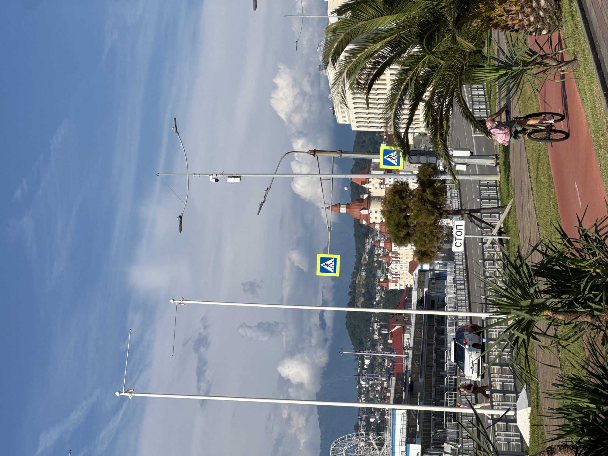 This sunny, eye-level outdoor shot captures a vibrant urban scene in Sirius, Russia, likely near the Olympic Park or Imeretinsky Resort. The sky above is predominantly blue with scattered white cumulus clouds, some appearing anvil-shaped, suggesting a warm day.
In the immediate foreground, lush green foliage and several palm trees with thick, patterned trunks line a red asphalt bicycle path, on which a person with a pink backpack cycles away from the viewer on a dark mountain bike. To the left of the bike path, another stretch of green grass leads to a sidewalk.
Separating the foreground from the midground is a paved road, barricaded by a series of low metal fences. On the road, a white car with red accents on its roof and side mirrors is partially visible, appearing to be at a standstill or moving slowly. Two pedestrians walk on the sidewalk parallel to the car. A white "СТОП" (STOP) sign is clearly visible on the right side of the road, mounted on a pole. Tall, white, cylindrical flagpoles with horizontal arms extend vertically through the frame on the left, and a multi-headed street light pole equipped with a surveillance camera stands prominently in the center-right midground. Two yellow square signs featuring a walking person icon (pedestrian crossing) are visible above the road.
The background showcases a developed urban landscape against a backdrop of distant, verdant mountains partially obscured by clouds. Distinctive red-roofed, castle-like buildings are scattered across the mid-distance, one with multiple conical spires. To the right of these, a large, modern white building with numerous arched windows is prominent. A large white Ferris wheel is partially visible on the far left, suggesting a recreational area or amusement park.
