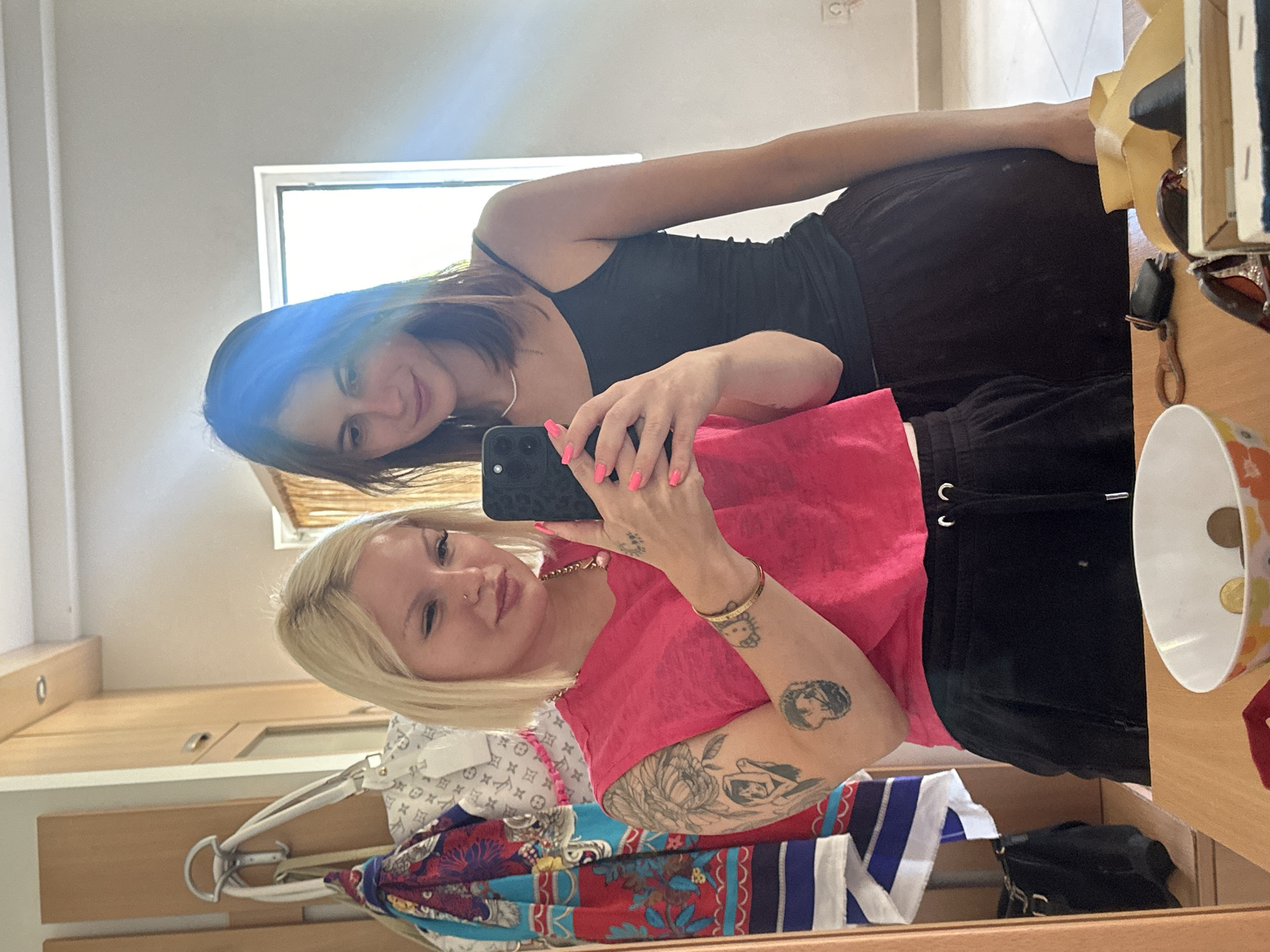 This is a medium shot, indoor, mirror selfie of two young women in what appears to be a residential setting in Bečići, Montenegro. The woman on the left, with blonde hair and wearing a bright pink tank top and black sweatpants, is holding up a smartphone to take the picture. She has tattoos on her left arm and is wearing a necklace and bracelet. Her nails are painted bright pink. The woman on the right, with brown hair and wearing a black tank top and dark sweatpants, stands slightly behind her, looking down at the phone. The background shows a closet or wardrobe with clothes hanging on the left, and a window with blinds on the right. A dresser or table is in the foreground, with a bowl containing coins, keys, sunglasses, and other small items. The lighting suggests it is daytime.