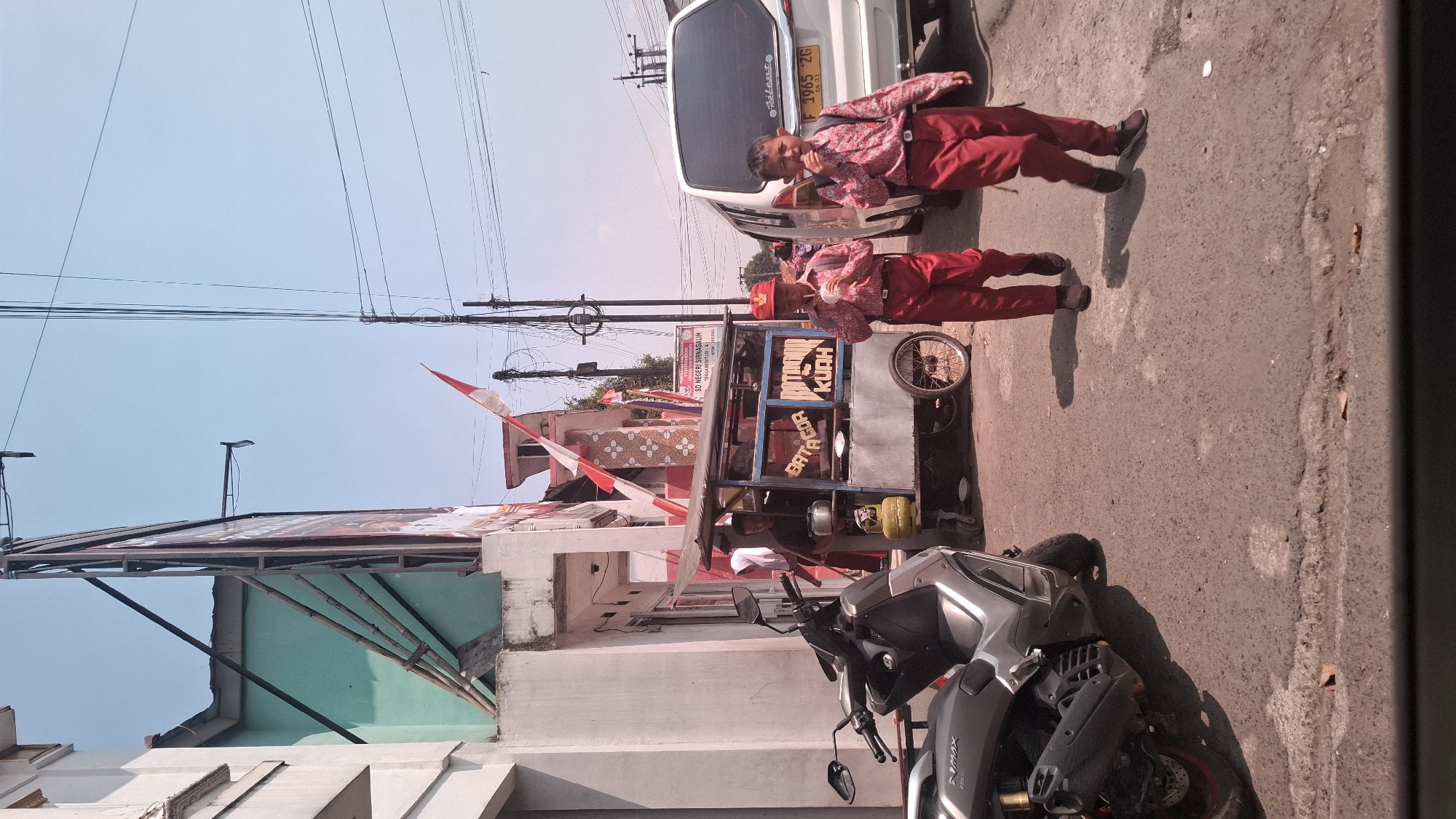 In the city of Sirnagalih, Indonesia, a street scene unfolds under a clear blue sky. Two young boys, dressed in matching red school uniforms with white shirts and red pants, walk on the right side of the frame. The boy on the left, wearing a red cap, holds a white cup to his mouth, while the boy on the right also appears to be holding something to his mouth. They are walking away from a food cart that is parked on the left side of the street. The cart is labeled "BATACOR KUAH" and has a yellow gas tank attached. A man, also in a dark shirt, is visible behind the cart. A gray motorcycle is parked to the left of the food cart. In the background, a white van is parked on the right side of the street, with a license plate that reads "F 1965 ZG". Buildings line the left side of the street, with a prominent billboard and an Indonesian flag displayed. Power lines crisscross the sky. The ground is paved with asphalt.