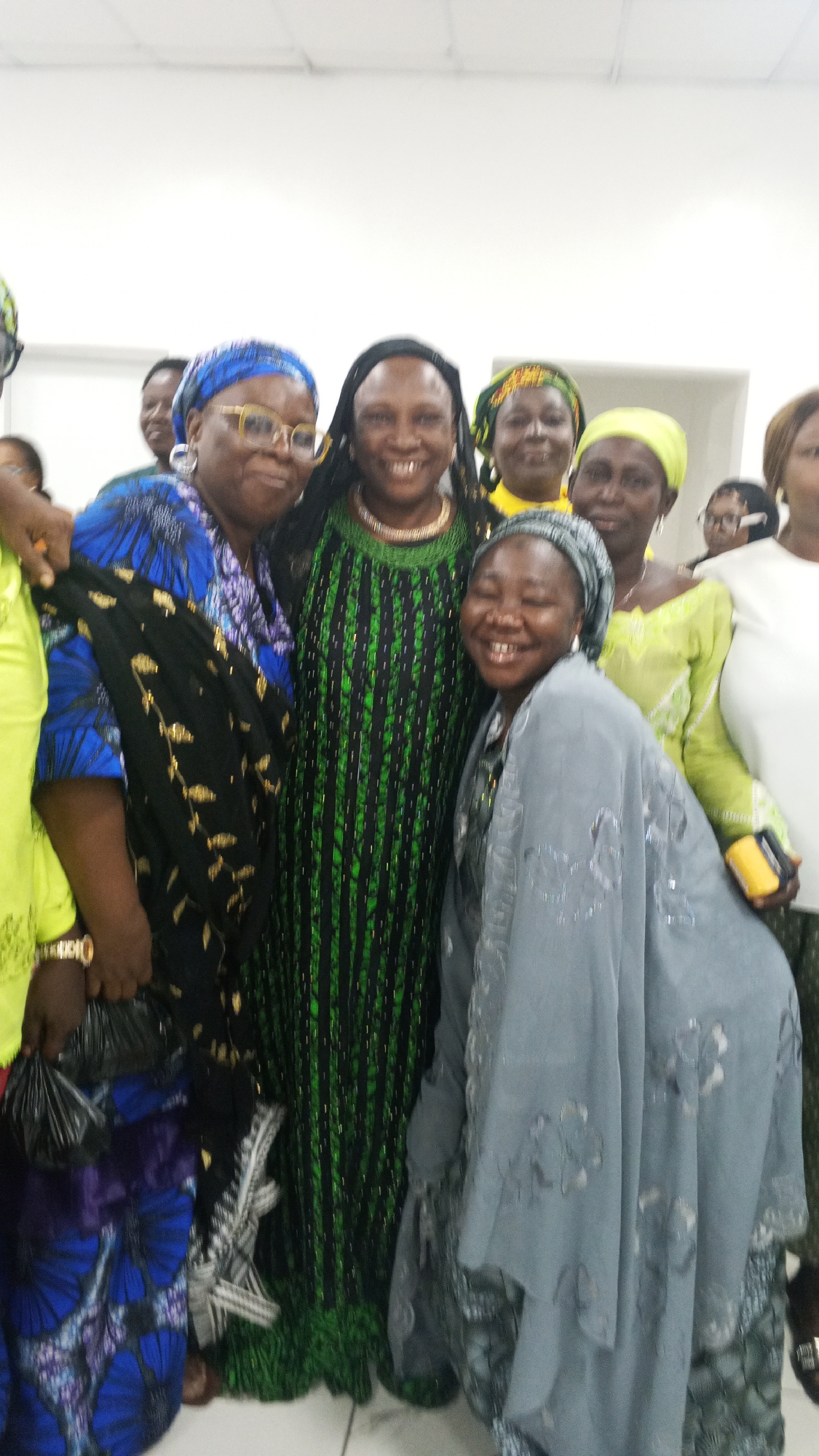 A joyful group of approximately ten to twelve women is captured in an indoor group photograph, likely taken in Lokoja, Nigeria. The central focus is on five women in the foreground, all exhibiting warm smiles and a sense of camaraderie.
On the left, a woman wearing a blue and purple patterned top and a blue headwrap, adorned with glasses and large hoop earrings, smiles broadly. Beside her, a woman in a long, black and green vertically striped dress with metallic accents and a black headwrap smiles widely at the camera, wearing a gold-colored necklace. To their right, another woman kneels or bends forward, her face radiant with a joyous, eyes-closed smile, dressed in a grey headwrap and a matching patterned top or shawl. Partially visible on the far left, a woman in a bright yellow top displays a gold watch and holds a black plastic bag, while on the far right, another woman in a white top holds a yellow object, possibly a phone or a small bag.
Several other women are visible in the background, also smiling and wearing headwraps in various colors like yellow and green, further contributing to the celebratory atmosphere. The women are dressed in colorful, traditional attire. The setting is a brightly lit indoor space with plain white walls, suggesting a social gathering, community event, or celebration. The overall mood of the image is one of happiness, unity, and shared enjoyment. No discernible text is visible in the photograph.