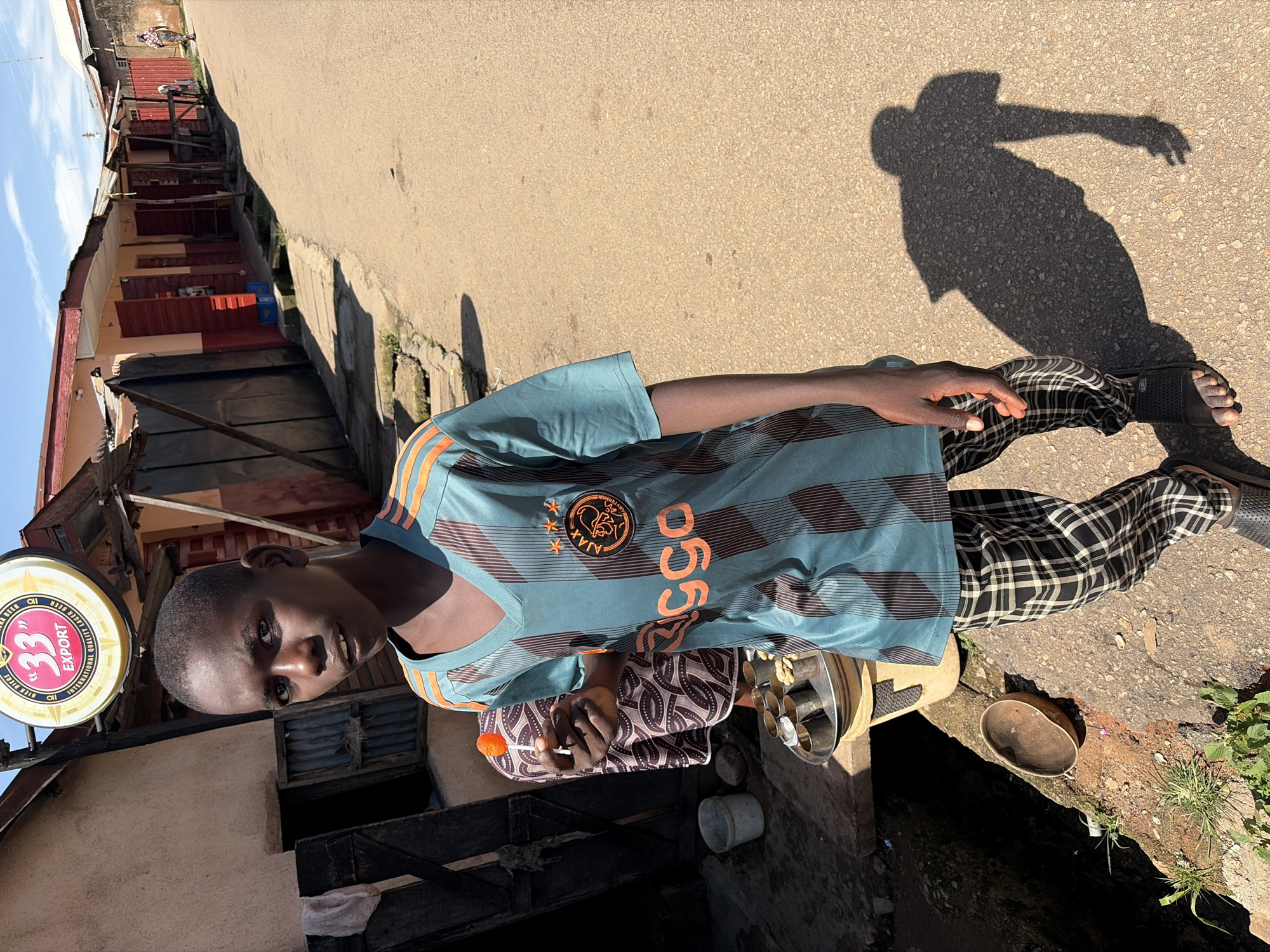 The image shows a young Black boy standing on a dirt road in an urban setting, likely in Jos, Nigeria, based on the context of the location. He is wearing a teal and orange striped t-shirt with an Ajax Amsterdam logo and checkered pants. In his right hand, he holds an orange lollipop. To his left, there's a stall or shop with a circular sign that reads "33 EXPORT". Behind him, a woman in a patterned dress stands near a tray with small metal cups. The environment appears to be a street lined with small businesses, some with metal shutters. The ground is dusty and uneven, with some debris and vegetation visible along the roadside. The lighting suggests it is daytime, with a clear sky. A shadow of the boy is cast on the road to his right, indicating the sun's position.