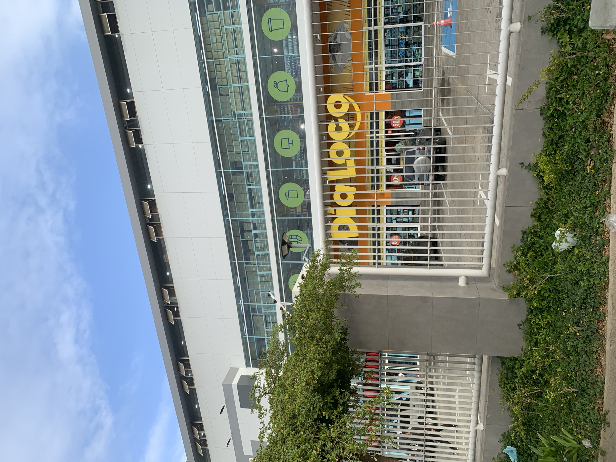 An exterior shot captures a modern commercial building, likely a store, under a bright blue sky with scattered white clouds. The building's facade is characterized by white panels and large glass windows. A prominent yellow sign, reading "Dia Loco" in a stylized font, is displayed on an orange background, indicating the establishment's name.
Through the large windows of the upper floors, rows of stacked boxes are visible, suggesting storage or inventory. Green circular icons, depicting various household items or product categories such as a cup, a pitcher, and what looks like an appliance, are affixed to the glass facade.
In the foreground, a white metal fence spans horizontally across the image, partially obscuring the entrance area of the store. Behind the fence, a small, dark-colored car is parked, and part of an orange traffic cone is visible. Lush green foliage, including bushes and a small tree, grows along the perimeter of the building and near the fence.
Inside the store, visible through the fence and windows, red signs with "50%" indicate ongoing sales. The overall scene suggests a commercial area during the daytime. The location is Porlamar, Venezuela.