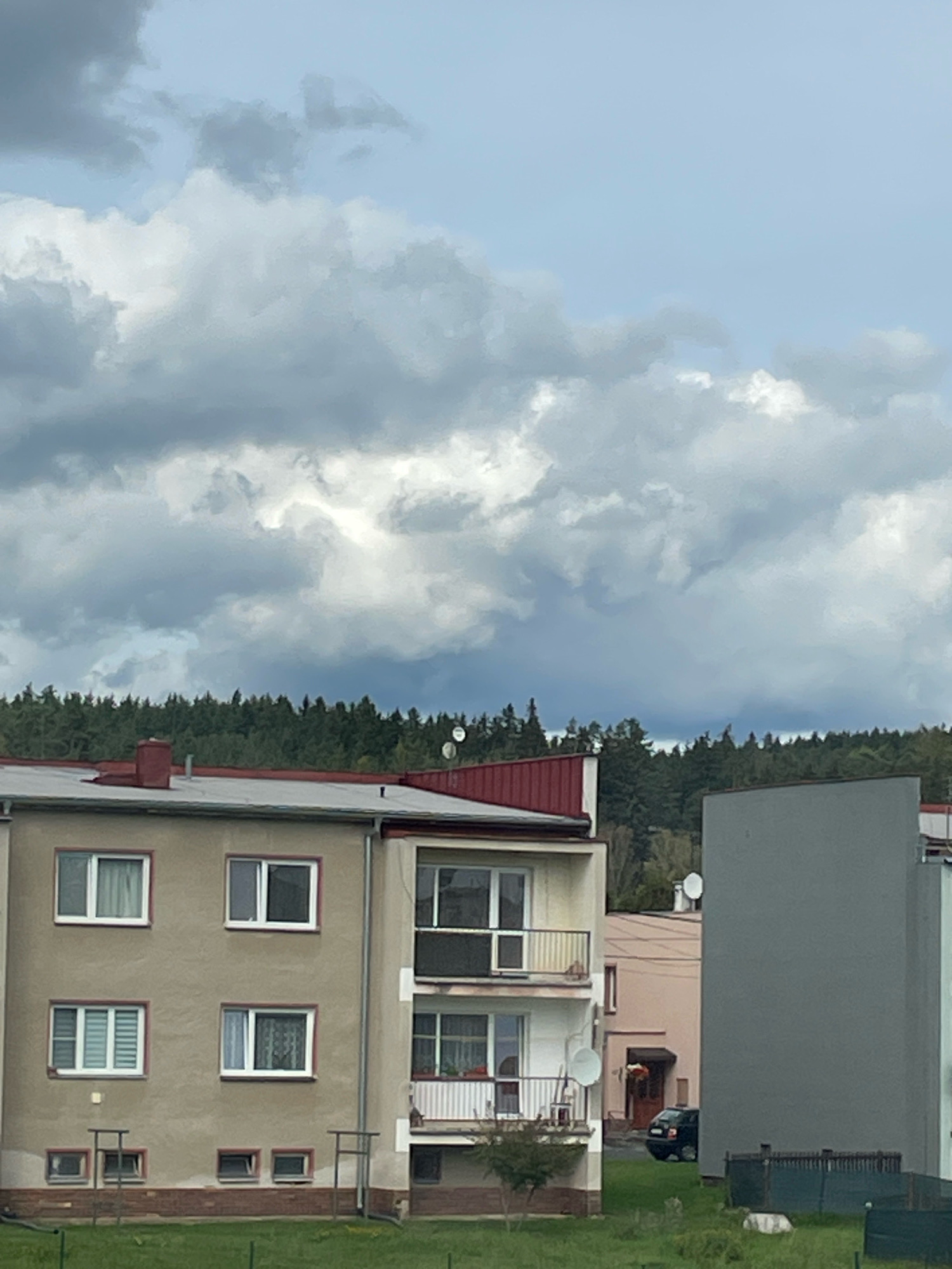 The image captures a view of buildings in the city of Starý Rybník, Czechia, under a cloudy sky. In the foreground, a portion of an apartment building is visible, characterized by its beige facade, white-framed windows, and a red-tiled roof with a chimney. To the right, another building with a large, plain gray facade is partially seen, and further to the right, a pinkish building with a dark car parked in front of it. Behind the buildings, a dense line of evergreen trees forms a natural backdrop. The sky is filled with dramatic, dark gray clouds, indicating overcast weather. The scene is devoid of people.