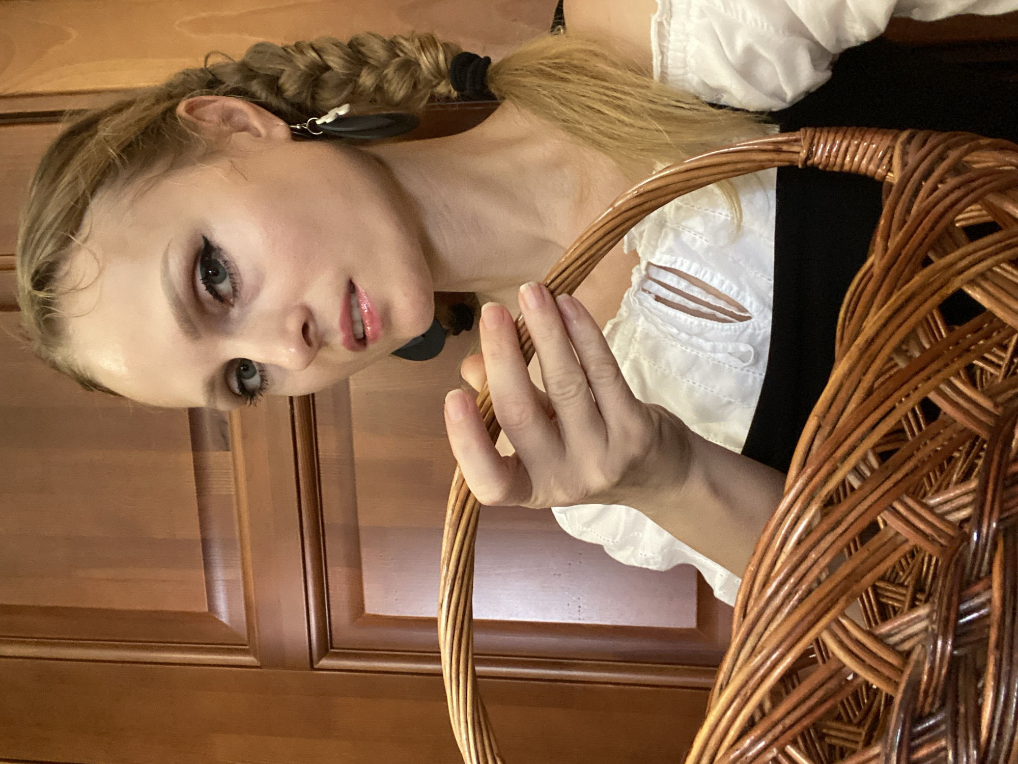 A woman with blonde hair in a braid and a white off-the-shoulder top with a black undergarment holds a woven basket. She is wearing large dark earrings. The background appears to be a wooden door. The image was taken in Lida, Belarus.