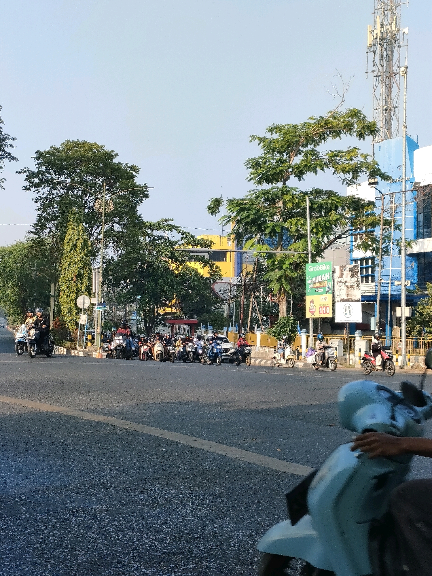 The image depicts a street scene in Banjarmasin, Indonesia, likely during daylight hours with a clear sky. In the foreground, a light blue scooter is partially visible, with a person's hand resting on its handlebars. The scooter is on a paved road with a yellow line marking the lane. In the middle ground, a busy intersection is filled with numerous scooters and motorcycles, many with riders. They appear to be stopped or waiting, suggesting traffic or a signal. Several tall trees are prominent in the background, some with dense foliage. Behind the trees, buildings can be seen, including a yellow building and a blue building with a large telecommunications tower on top. Advertisements are displayed on a pole near the intersection, one of which is for "GrabBike" with the word "MURAH" (cheap) visible. The overall impression is of an active urban environment with typical street life and transportation.
