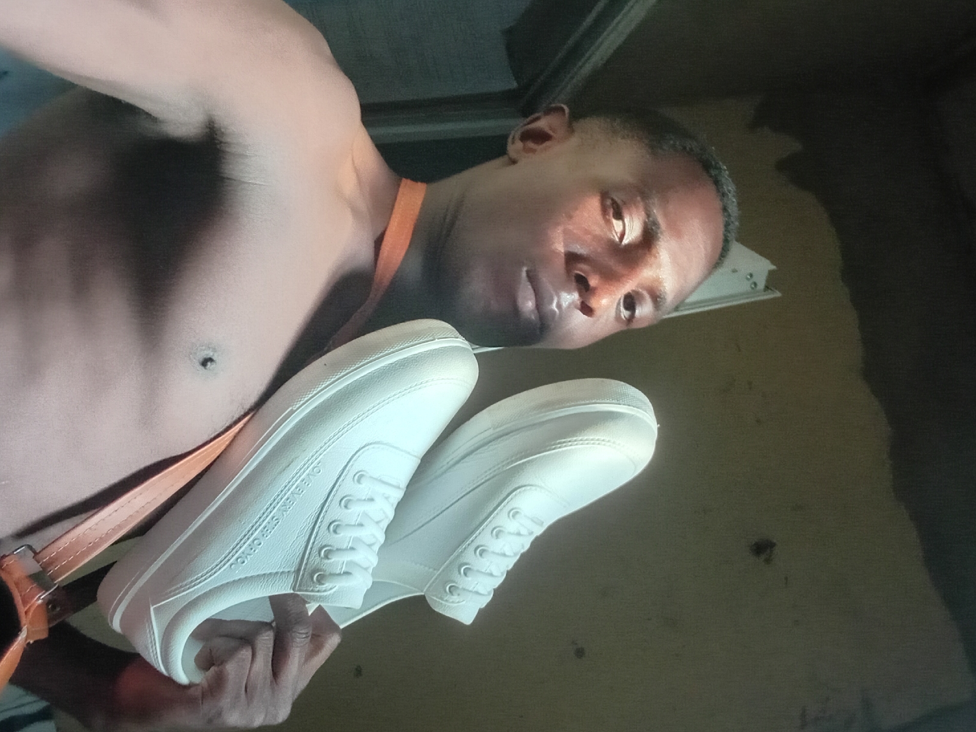 A shirtless, dark-skinned man is holding a pair of white sneakers in front of him. An orange strap crosses his chest. The background is out of focus but appears to be an interior space. There is no indication of the location being Akwete, Nigeria.