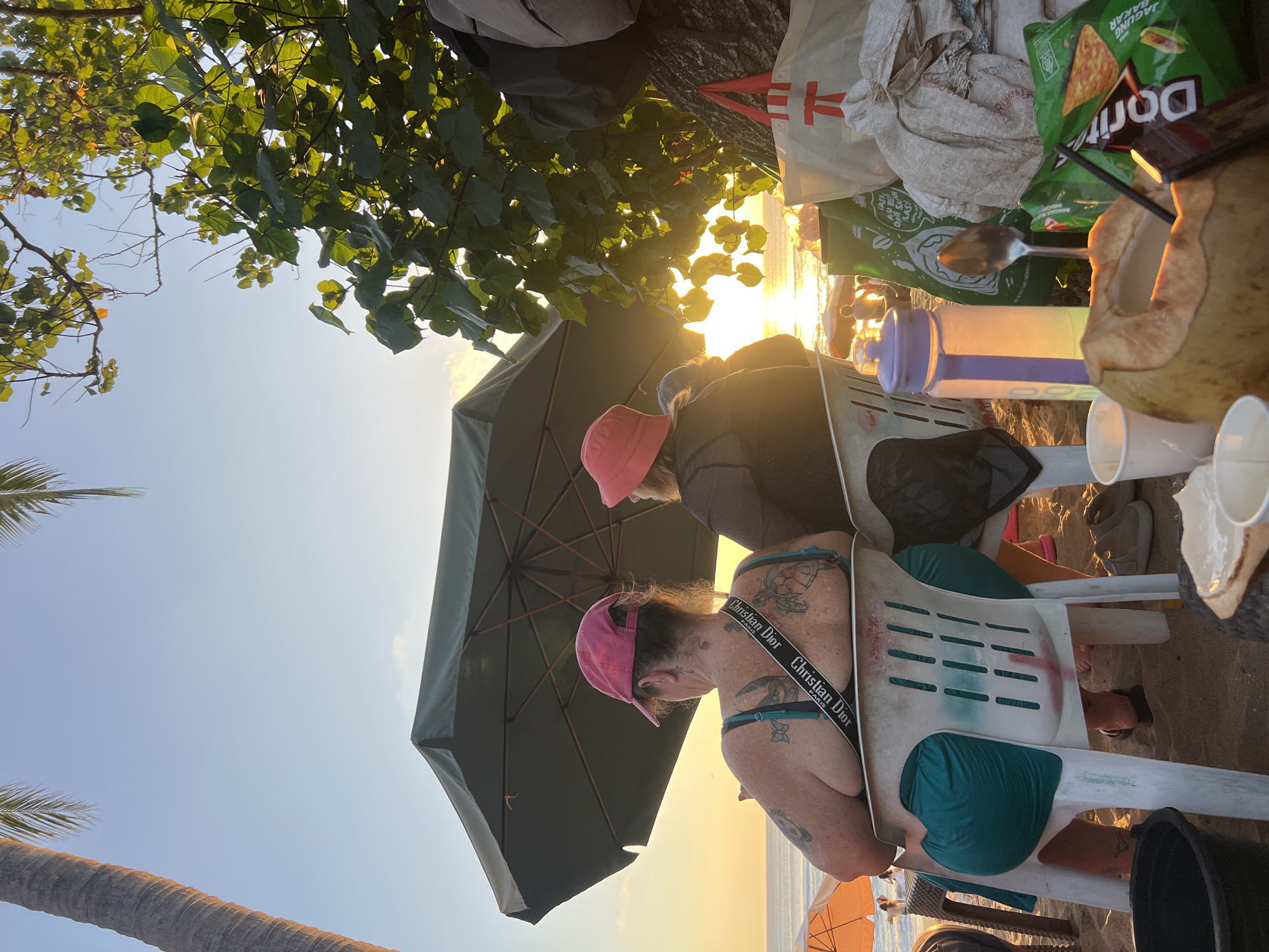 This image captures a beach scene in Kuta, Indonesia, during sunset. Two individuals are seated on white plastic chairs, facing away from the camera, under a large green umbrella. The person on the left wears a pink cap and a teal top, with visible tattoos on their back and arms. A bag with "Christian Dior" branding is slung over their shoulder. The person on the right wears a red hat and a dark top.
In the foreground, various items are placed on the sand, including a halved coconut with a straw, cups, a purple water bottle, a spoon, bags of chips, and other miscellaneous items. The ocean and a golden sunset are visible in the background, with palm trees and foliage framing the scene. The overall atmosphere suggests a relaxed, tropical evening on the beach.
