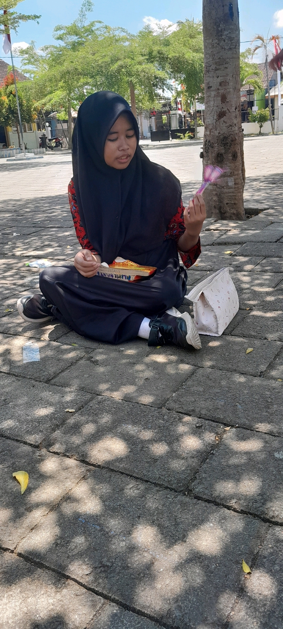 A young girl wearing a black hijab and a red and black patterned shirt is sitting cross-legged on a paved ground in a sunny outdoor area, possibly a schoolyard or park in Rogojampi, Indonesia. She is holding a small pink and purple toy in her right hand. To her left, resting on the ground, is a light-colored clutch bag with a subtle pattern. In front of her, on her lap, are two books, one yellow and one orange, with their titles partially visible. She is looking down at the books. The ground is made of interlocking rectangular concrete slabs, and dappled sunlight filters through the leaves of a nearby tree, casting shadows on the pavement. In the background, trees with lush green foliage are visible, along with a glimpse of buildings and a flagpole with a white and red Indonesian flag. The scene suggests a casual moment of rest or study outdoors.